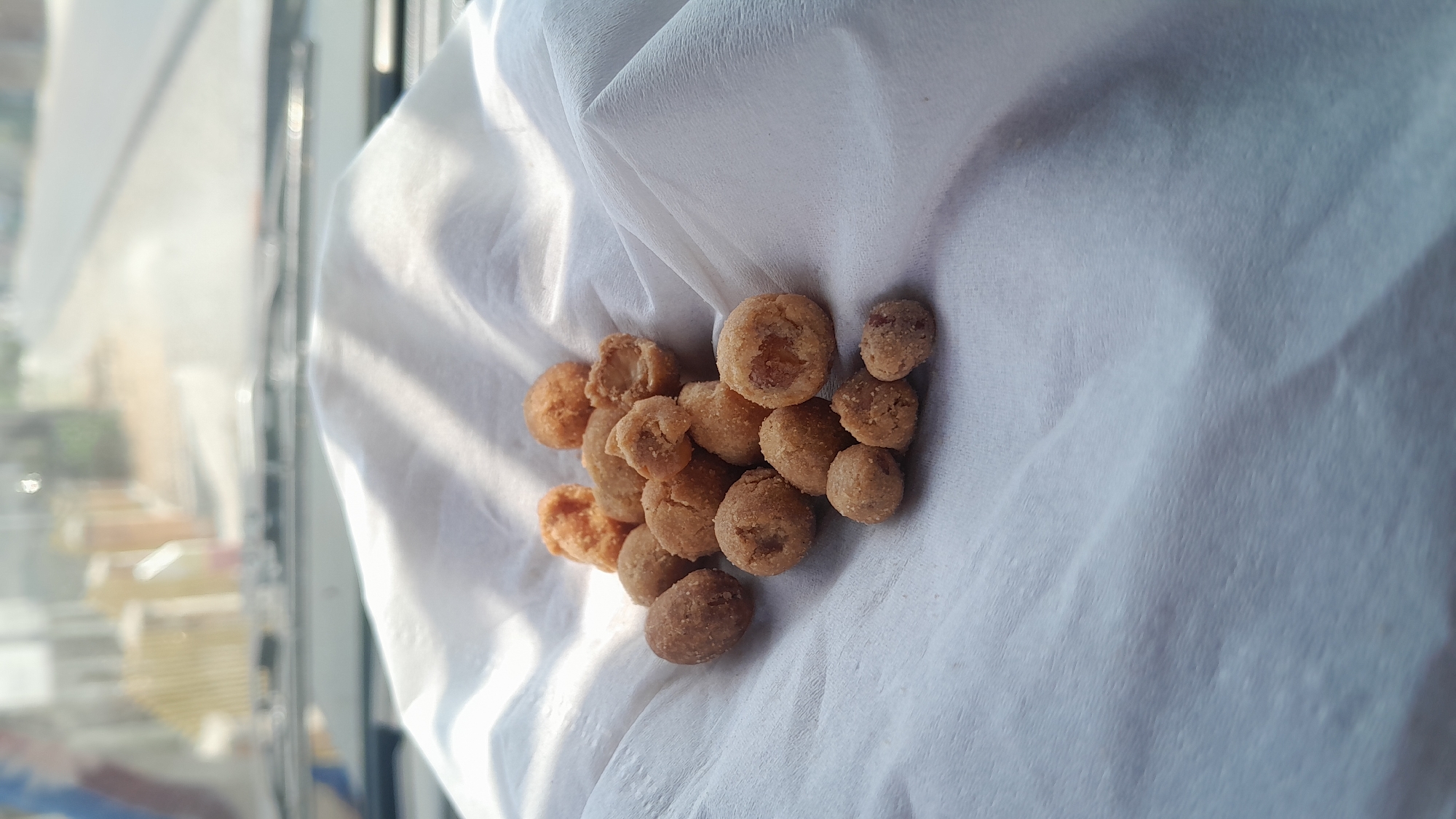 A pile of roasted, coated peanuts sits on a white, textured fabric. The peanuts are golden brown and appear to be of various sizes, with some having a crispy coating and others appearing more natural. The fabric is draped and folded, creating shadows and highlights that give it depth. In the blurred background, a street scene is visible, suggesting an outdoor setting, possibly from inside a vehicle due to the presence of what appears to be a windshield and dashboard. The overall lighting indicates it is daytime. The context of Bangkle, Indonesia, is not visually discernible from the provided image.