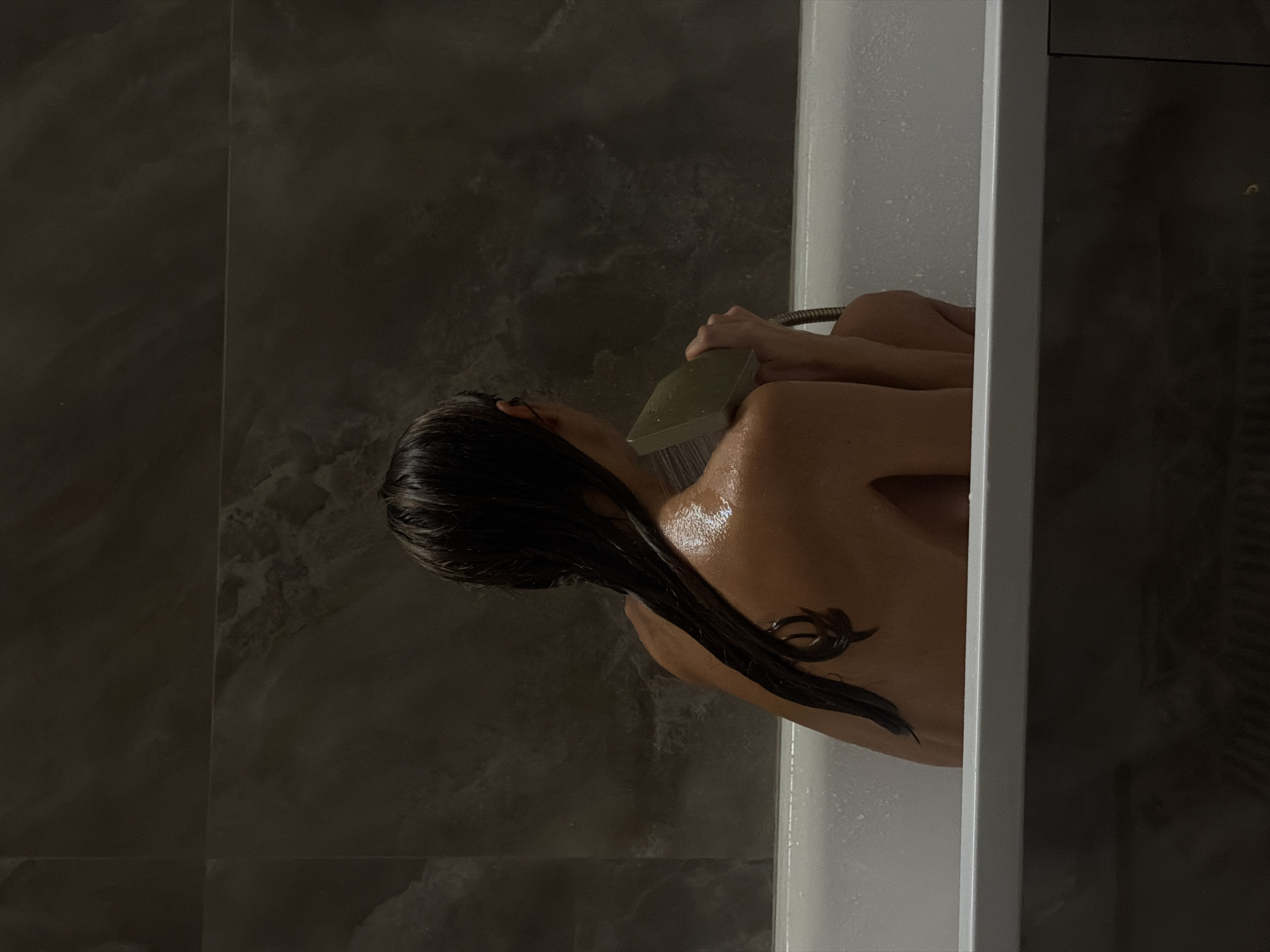 A person is in a white bathtub, holding a shower head over their shoulder and back. Their wet, dark hair is hanging down their back. The bathroom has dark, marbled walls. The location is unknown and cannot be verified as Krasnoyarsk, Russia based on the provided visual information.