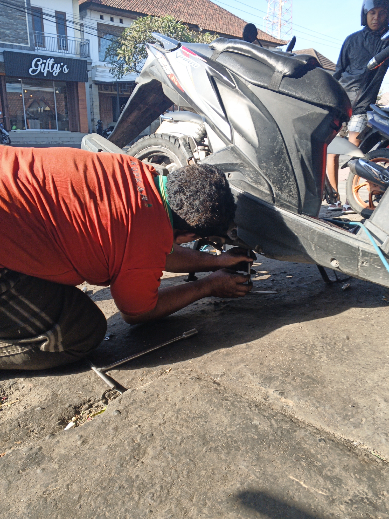 An outdoor scene in Sukawati, Indonesia, depicts a person kneeling to repair a black and silver motorcycle. The individual, wearing an orange t-shirt and dark pants, is using a silver tool to work on the underside of the vehicle. A second person, wearing a black jacket and plaid shorts and a helmet, stands nearby, observing. The background reveals a building with a sign reading "Gifty's" and a portion of a red-tiled roof, suggesting a commercial or residential area. The ground is made of cracked concrete and dirt. The lighting indicates a bright, sunny day.