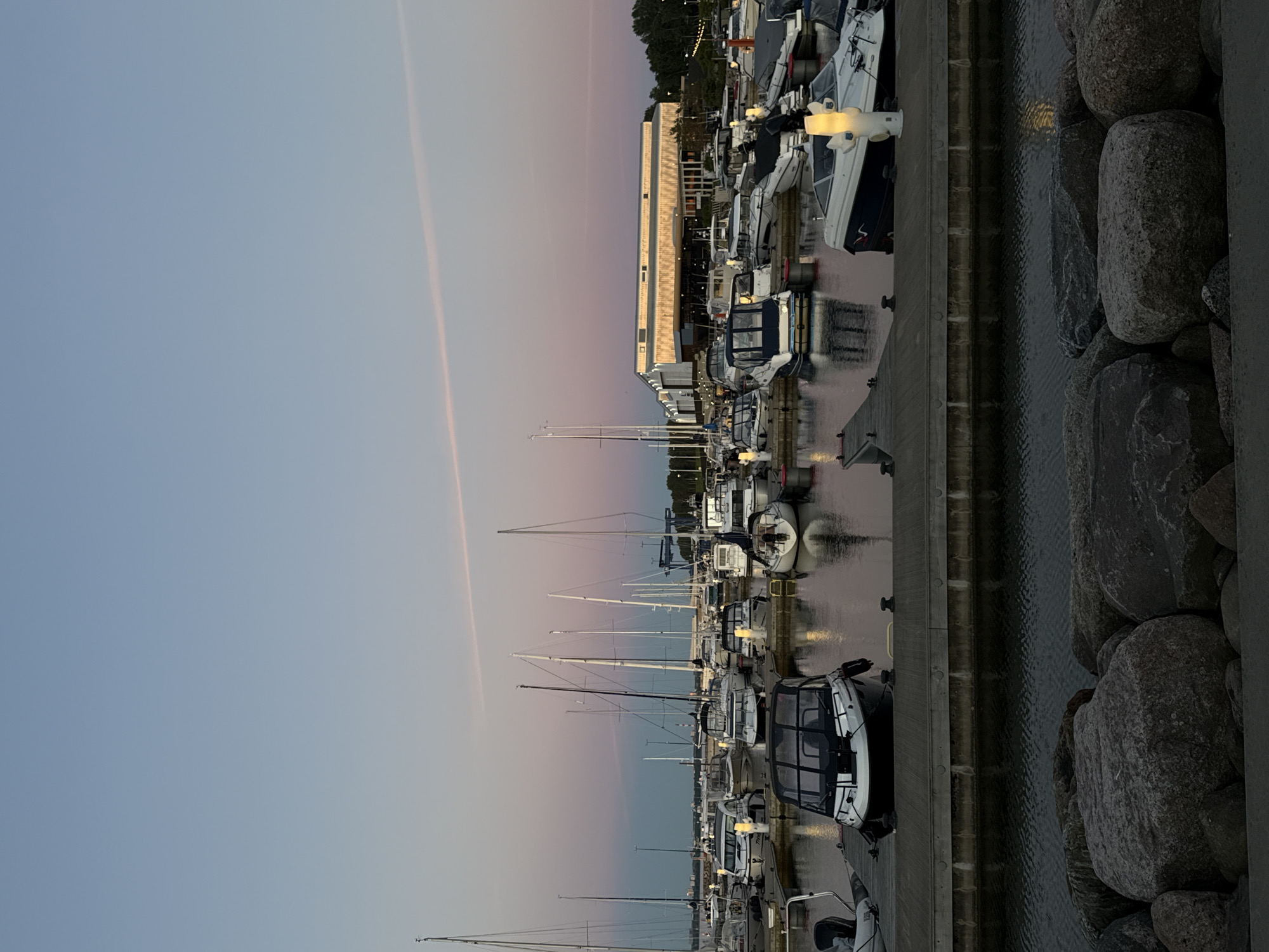 The image shows a marina at dusk in Tallinn, Estonia. A variety of boats, including sailboats and motorboats, are docked along a wooden pier. In the background, a modern building with large windows is visible, illuminated by interior lights. The sky is a gradient of pale blue and pink, with a thin, wispy cloud streak. The water in the marina reflects the lights of the boats and the building. In the foreground, large rocks are piled along the edge of the water. No people are visible in the image.