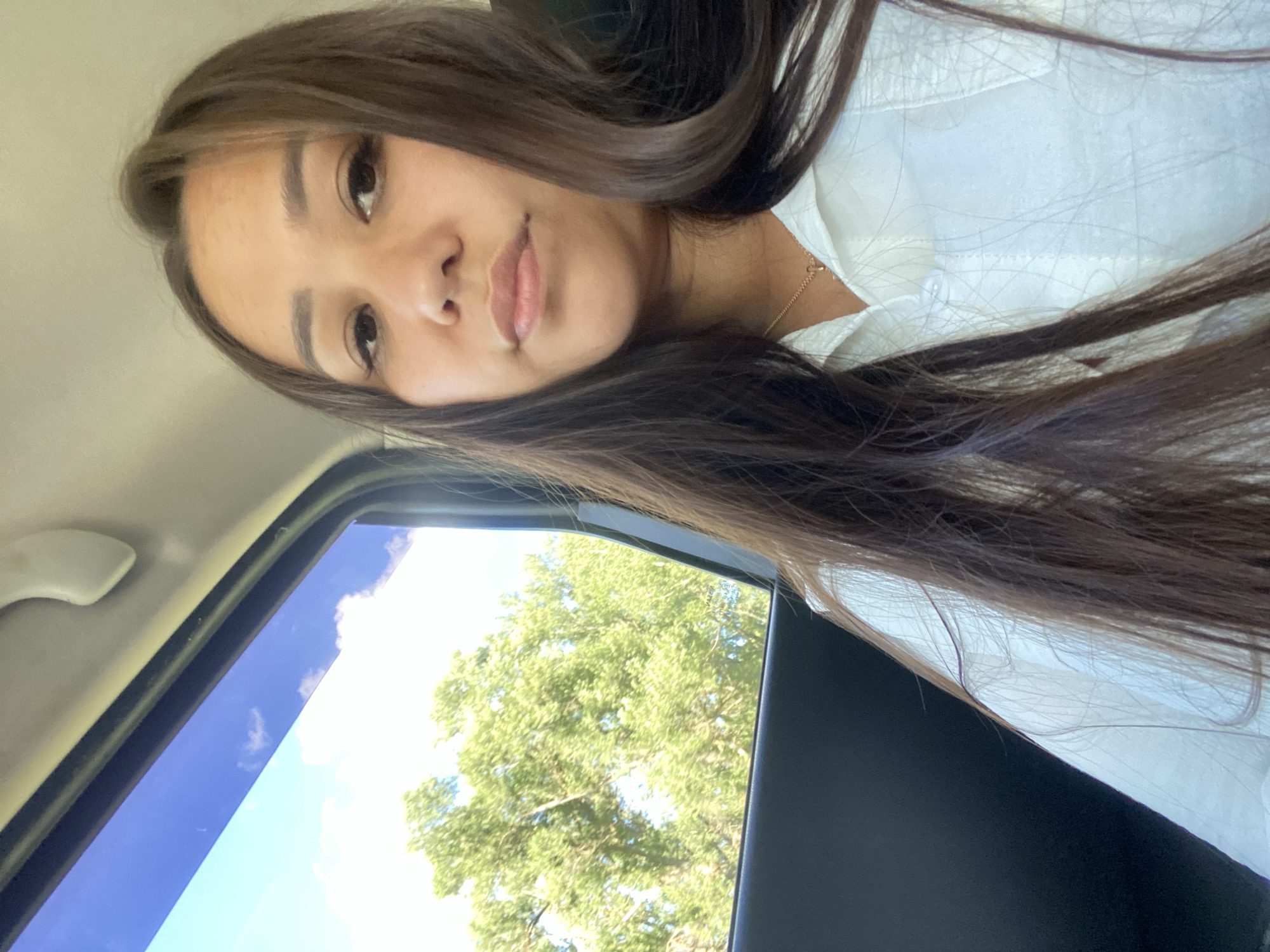 A young woman with long, dark brown hair is captured in a selfie from inside a vehicle. She is wearing a white collared shirt and a delicate gold necklace. Through the car window to her left, lush green trees and a bright blue sky with scattered clouds are visible. The interior of the car appears to be dark-colored, with a white handle visible at the top of the frame. The scene suggests a travel setting, possibly during the daytime.