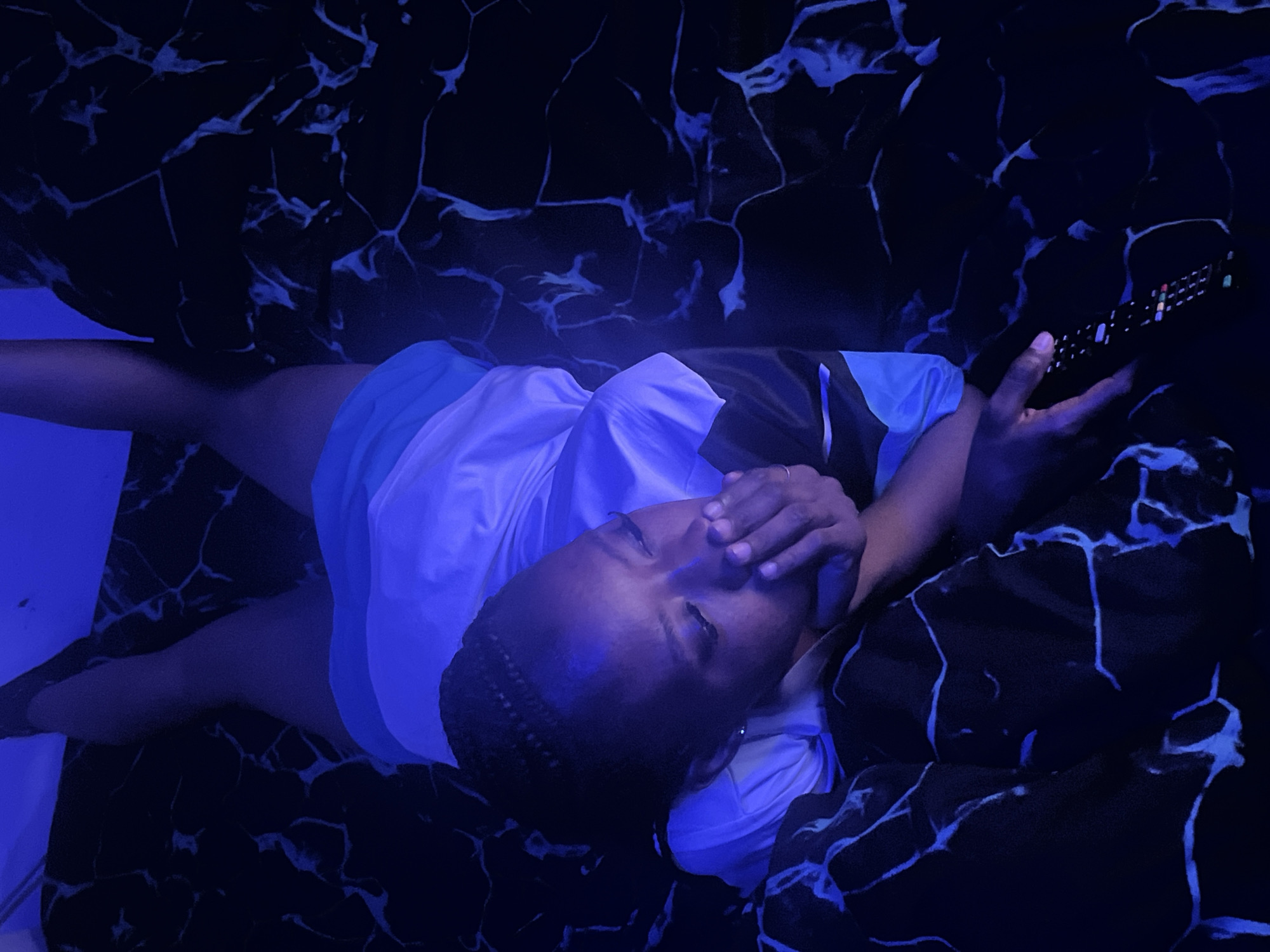 The image displays a person lying down in a dimly lit indoor setting, heavily saturated with a blue light, likely during the night in Jos, Nigeria. The individual is positioned diagonally across the frame, viewed from above, with their head slightly turned towards the viewer. One hand is raised, partially covering their face, with a simple ring visible on a finger.
The person has braided hair and is dressed in a light-colored t-shirt, possibly white with blue trim or accents, and what appear to be shorts. In their other hand, they hold a black remote control with illuminated, multicolored buttons, suggesting they are interacting with an electronic device, such as a television.
The surrounding environment consists of a dark fabric, likely upholstery of a couch or bedding, featuring a striking black background with glowing blue-white patterns resembling cracked ice or abstract veins, which are vividly highlighted by the blue light. The dominant blue illumination bathes the entire scene, coloring the person's skin and clothing with cool tones and creating a distinct, somewhat mysterious atmosphere. There is no visible text in the image.