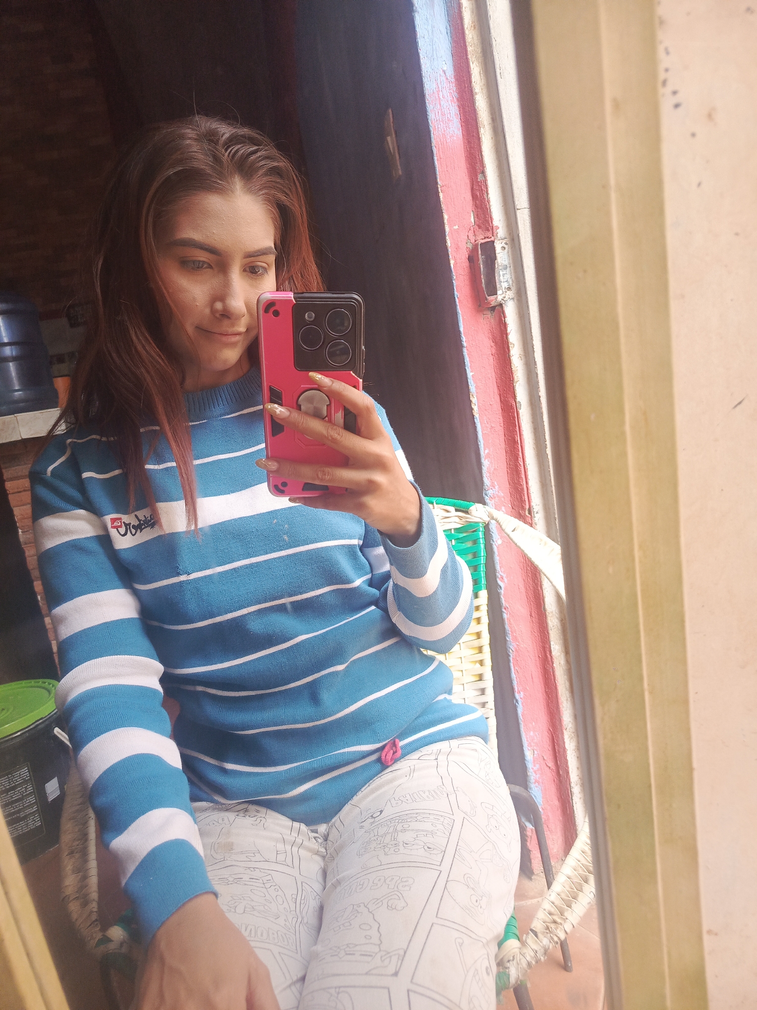 A young woman takes a selfie with a pink-cased smartphone in front of a mirror. She has reddish-brown hair and is wearing a blue and white striped sweater and white comic-print pants. She is seated in a chair with woven elements and what looks like a small green bucket to her left. Behind her is a dark indoor space with a large blue container. The window frame next to her is painted red and blue. The location is likely a residential area, inferred from the casual clothing and domestic setting, but without further information, a precise location in Juan Griego, Venezuela cannot be determined.