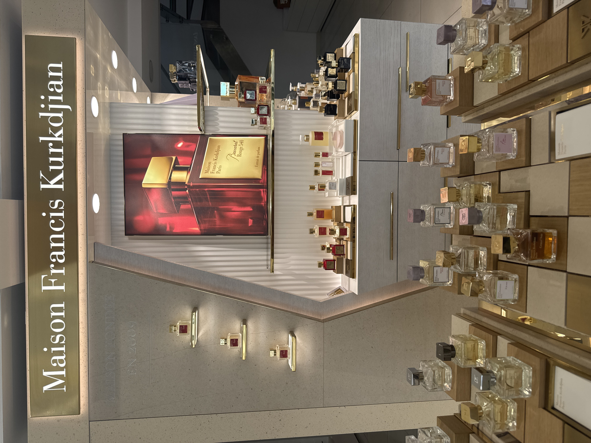 This is a medium shot, eye-level, interior photograph of a Maison Francis Kurkdjian fragrance store display. The store is located in Germasogeia Municipality, Cyprus. The display features multiple bottles of perfume on tiered shelves and stands, illuminated by spotlights and ambient lighting. The brand name "Maison Francis Kurkdjian" is prominently displayed in large, illuminated white lettering on a gold sign above the display.
In the background, a large promotional image of a red and gold perfume bottle is visible. The main display area includes several shelves mounted on a textured, light-colored wall, each holding a single bottle of perfume. Below this, a white wooden cabinet with gold drawer pulls serves as the base for further displays.
The foreground of the image showcases an array of perfume bottles arranged on wooden stands and within clear acrylic holders. The bottles are of various sizes and shapes, with some containing clear or amber-colored liquid, and others topped with gold or metallic caps. The lighting creates a warm and inviting atmosphere, highlighting the luxury products. There are no people visible in the image. The overall impression is one of a high-end retail environment specializing in designer fragrances.
