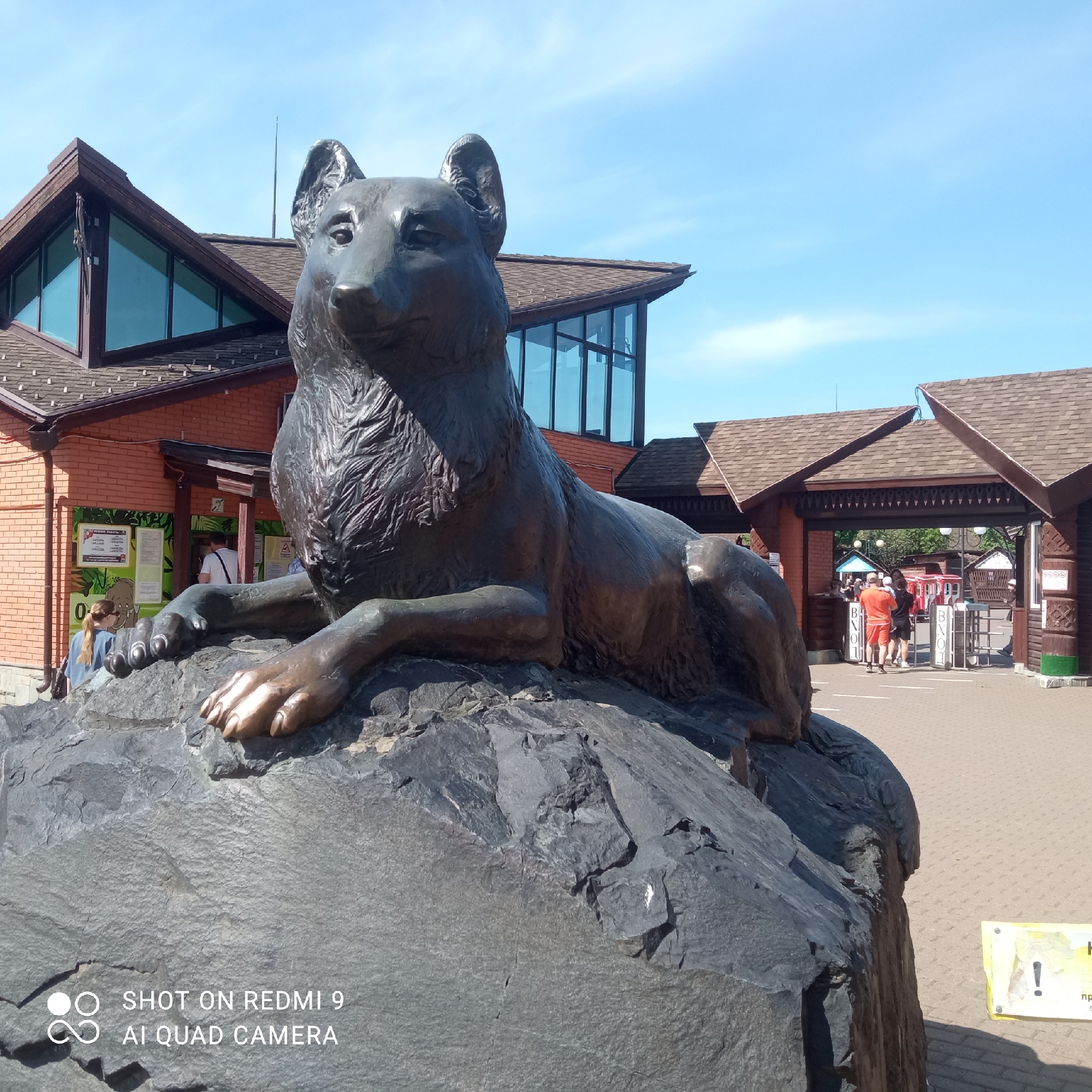 The image shows a bronze-colored wolf statue reclining on a large, dark gray rock. The statue is positioned in front of a building constructed of red brick and featuring a brown-tiled roof with sharp triangular gables. The building also has modern, geometric windows. In the background, a wooden archway with turnstiles marks an entrance area, possibly to a park or zoo. People are visible passing through the turnstiles, dressed in casual summer attire. The ground is paved with interlocking bricks. The sky is clear and blue, suggesting a sunny day. In the bottom left of the image is text indicating the photo was taken with a Redmi 9 AI Quad Camera. It is located in the city of Izhevsk, Russia.