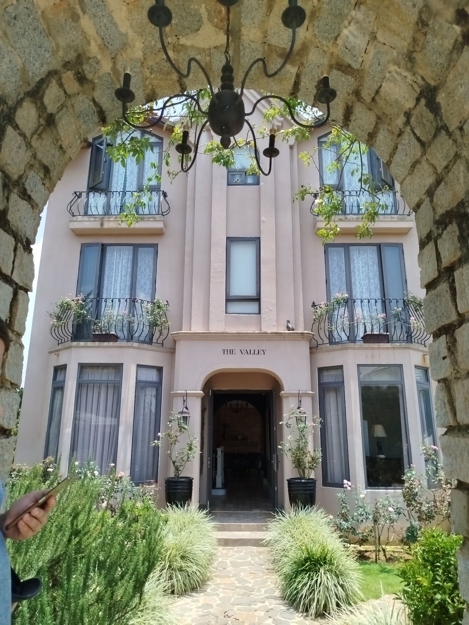 The image presents a view of a multi-story building framed by a stone archway. The building is beige-colored with dark-framed windows and small balconies featuring black iron railings. Some balconies have flower pots on them. A chandelier hangs in front of the building, with vines or climbing plants intertwined in its design. The entrance to the building is marked with "THE VALLEY" written above the doorway. Plants and bushes surround the base of the building along a paved walkway leading to the entrance. The scene appears to be outdoors, under daylight conditions. The location is Lang Biang - Đà Lạt, Vietnam.