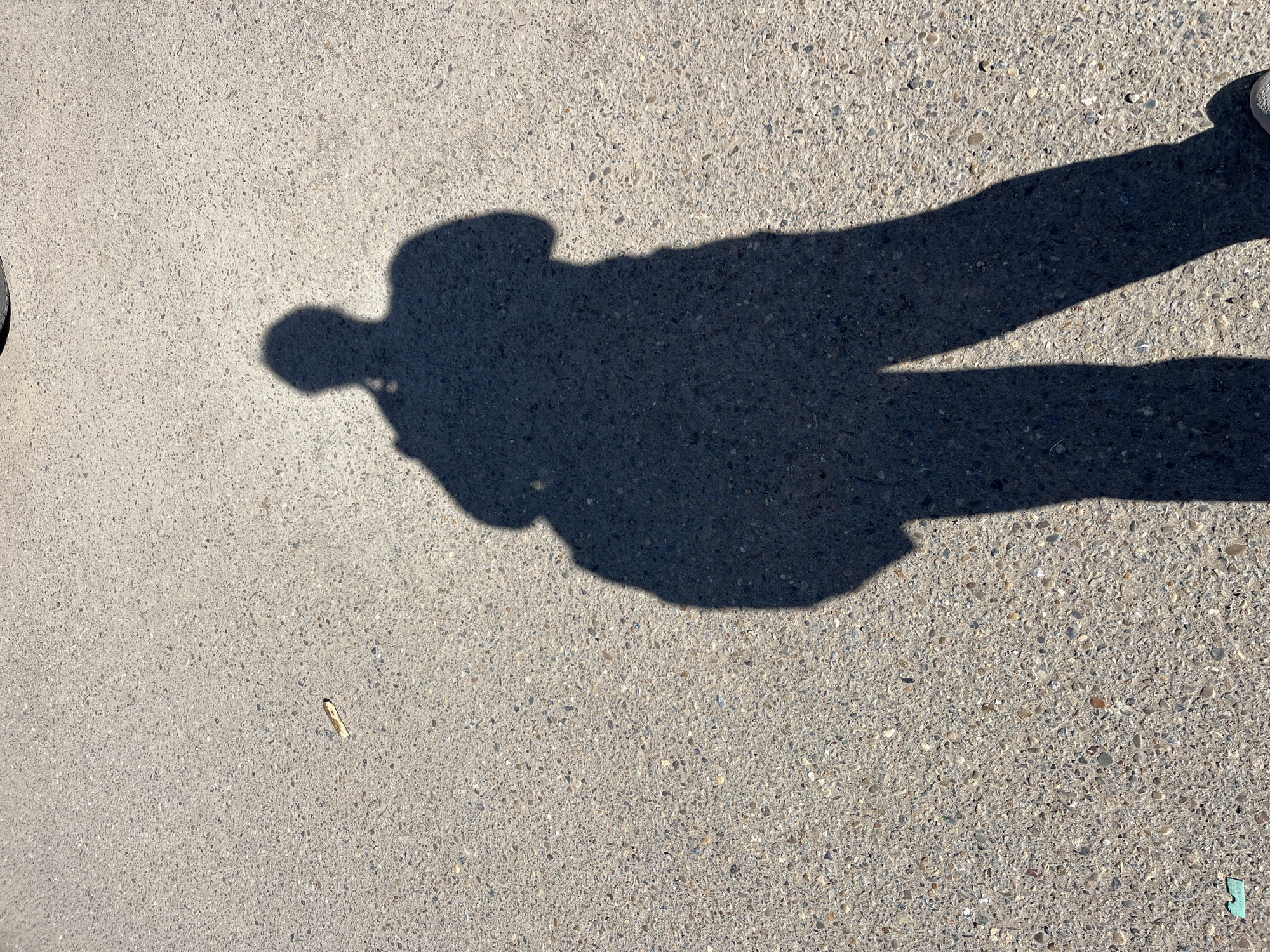 The image shows a person's shadow on a gray concrete surface. The shadow suggests the person is wearing a hat or has a rounded head, and is casting a shadow of their arms and legs. The concrete surface has a speckled texture. A small, light-colored object is visible near the lower left corner of the image. Part of a shoe or foot is seen in the upper right corner of the frame. The scene appears to be outdoors in daylight. Location cannot be determined.