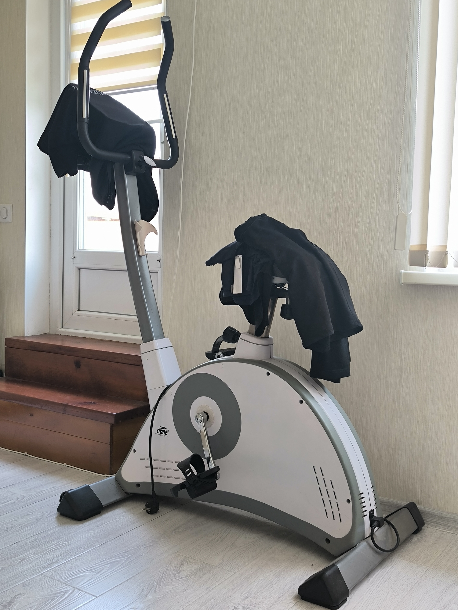 An exercise bike is positioned on a light-colored wooden floor, adjacent to a set of dark wooden stairs that lead to a doorway with a window. The exercise bike is predominantly white and gray, with black handlebars and pedals. Black clothing is draped over the handlebars and the central column of the bike. The background consists of a textured, light-colored wall and a window with horizontal blinds. The overall scene suggests an indoor setting, likely a room in a residential building. There is no indication of the specific location within Chișinău, Moldova.
