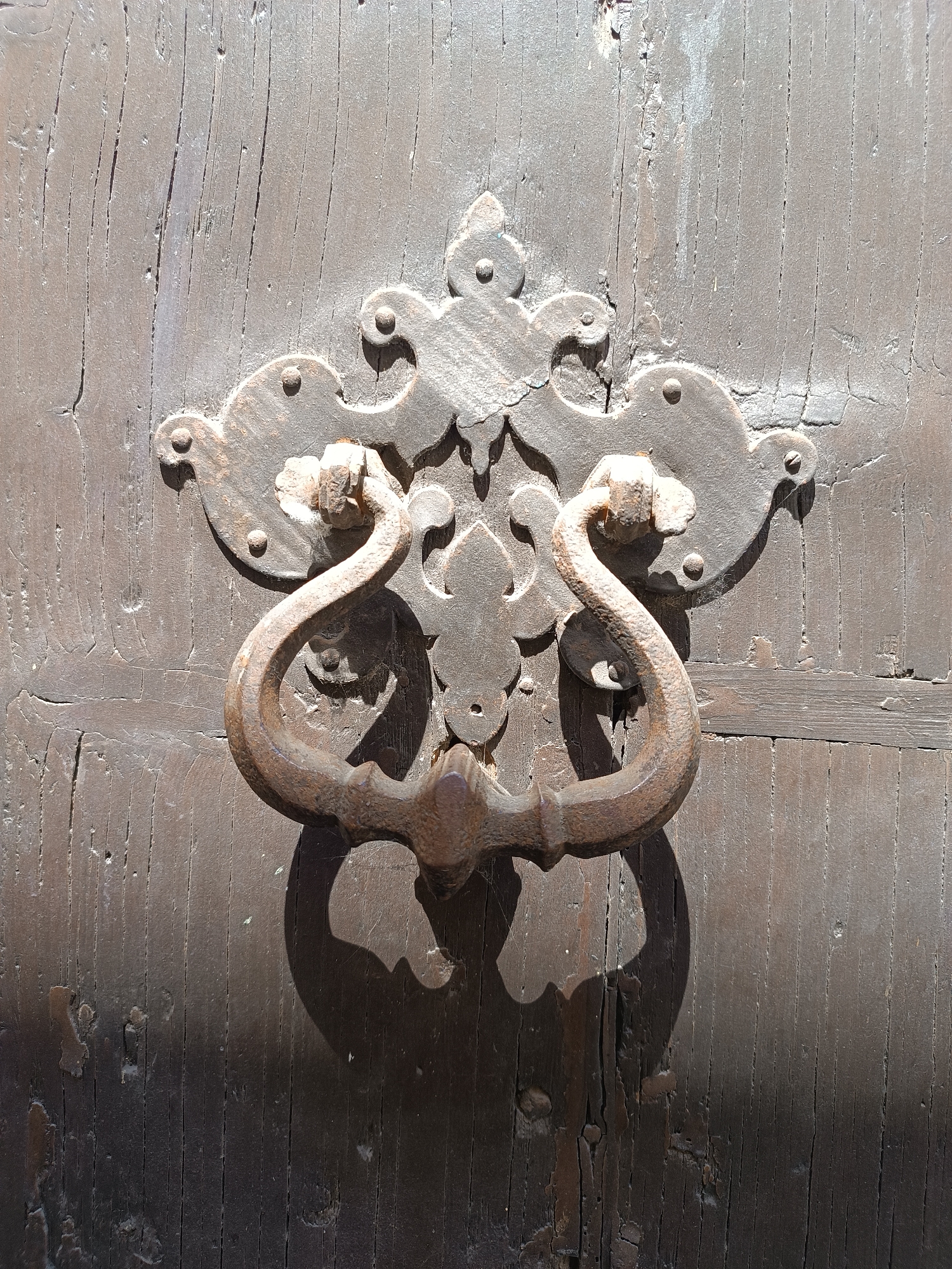 The image shows a close-up of a weathered, brown wooden door with an ornate iron door knocker. The knocker features a decorative backplate with floral and geometric elements, and a curved handle that casts a shadow on the lower part of the door. The door itself is aged and shows signs of wear, with visible wood grain and cracks. The image was taken in Mezzana Sant'Antonio, Italy.