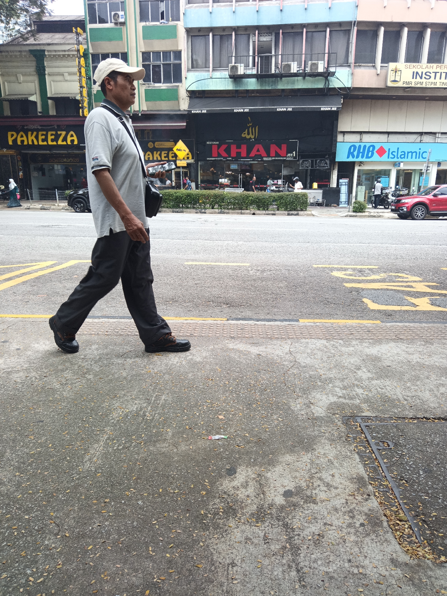 In Kuala Lumpur, Malaysia, a middle-aged man wearing a light-colored polo shirt, dark trousers, a white cap, and black shoes walks on a sidewalk next to a street. He carries a black shoulder bag. The sidewalk is made of concrete and is adjacent to a road marked with yellow lines. In the background, several buildings are visible. One building features a large sign with yellow lettering that reads "PA জীবাEEZA," and another has the words "KHAN JEE" and "KHAN" in large red letters, with Arabic script above. A blue sign that says "RHB Islamic" is also visible. A red car is parked on the street. The scene appears to be a commercial area.