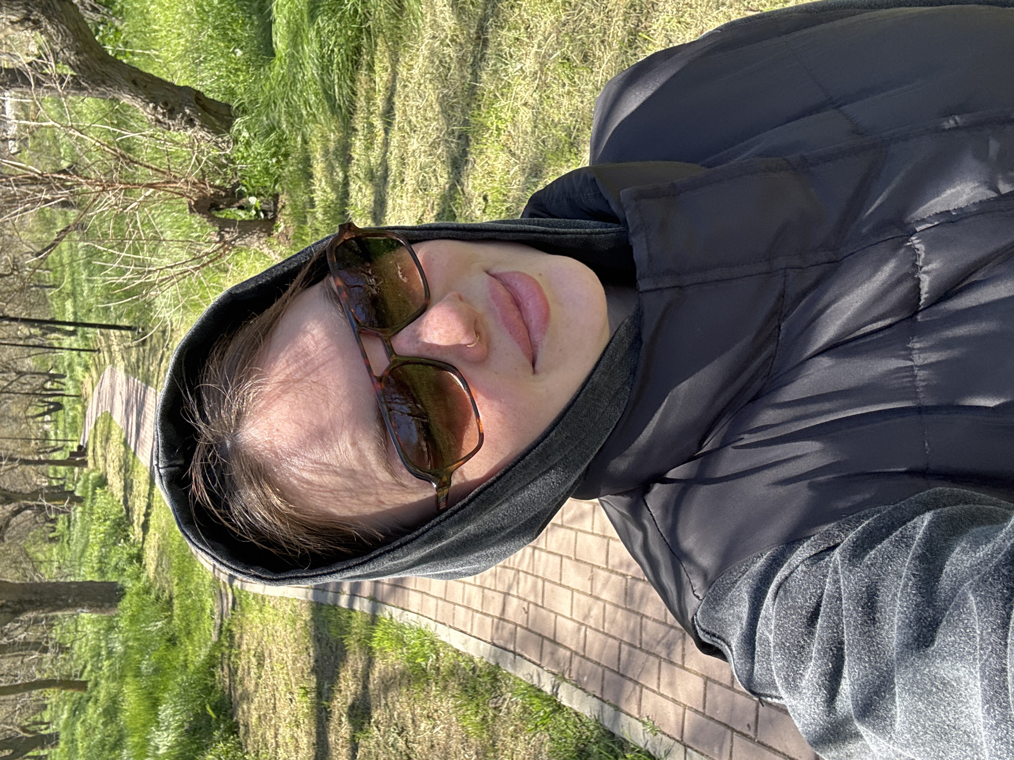 A person wearing a dark-colored hooded jacket and sunglasses takes a selfie on a brick pathway in what appears to be a park or green space. The location appears to be in Chornomorskoe, Ukraine, although the context comes from the image description, not the image itself. The person's face is visible, and they are looking slightly up towards the camera. The background shows green grass and trees. The sun is shining, casting shadows on the ground.