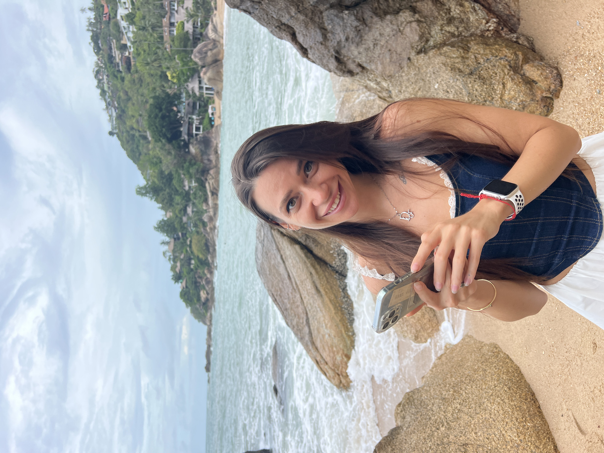 A woman smiles, holding a phone, standing on a beach in Baan Chaweng Noi, Thailand. She is wearing a blue denim top with white trim and a white skirt. She has an Apple Watch on her left wrist, with a red string bracelet alongside it. The beach is sandy, with large rocks bordering the water. The water is light green, and waves lap at the shore. Across the water, a hillside is visible, covered in lush green vegetation and houses. The sky is overcast.