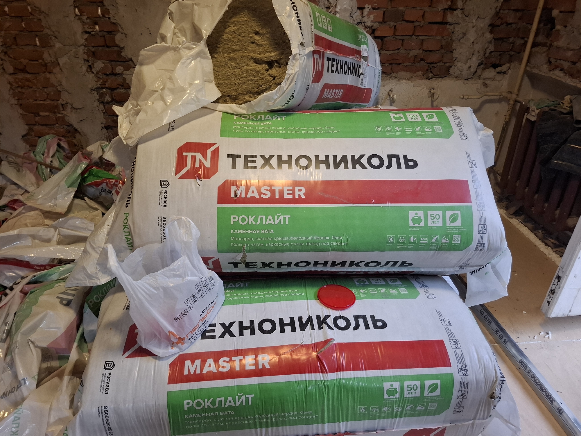 The image shows a storage area with several large rolls of insulation material stacked on top of each other. The rolls are wrapped in white and green plastic packaging with Russian text and branding that appears to be "ТЕХНОНИКОЛЬ" and "РОКЛАЙТ". Some of the rolls are partially opened, revealing the fibrous insulation material inside. In the foreground, there are also several plastic bags, one of which is open and empty. The background consists of a brick wall and a wooden floor. The overall impression is one of a construction or storage site. No people are visible in the image. The location is specified as Ryazan, Russia.