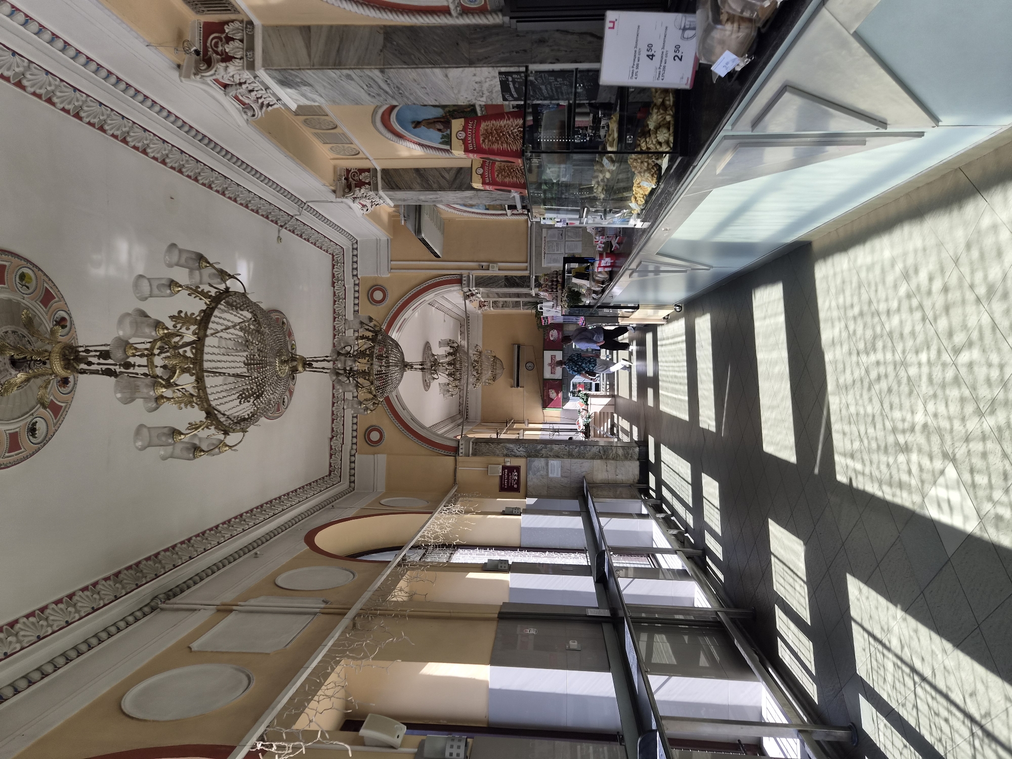 The image depicts the interior of a cafe or dining establishment, likely a self-service canteen, within Minsk, Belarus. The architecture suggests a historical or grand building, with ornate chandeliers hanging from a high ceiling adorned with decorative moldings and painted medallions. The walls are painted a pale yellow, with arched doorways and windows. Sunlight streams in from the left, casting geometric shadows across the tiled floor, creating a visually dynamic scene.
On the right side of the image, a long counter displays various baked goods in glass cases. Several customers are visible, including two individuals walking away from the camera down the hallway on the left, and another person or two near the counter on the right. The counter area appears to be a point of sale, with signage and pricing visible. The overall atmosphere is one of a functional but aesthetically pleasing public space.