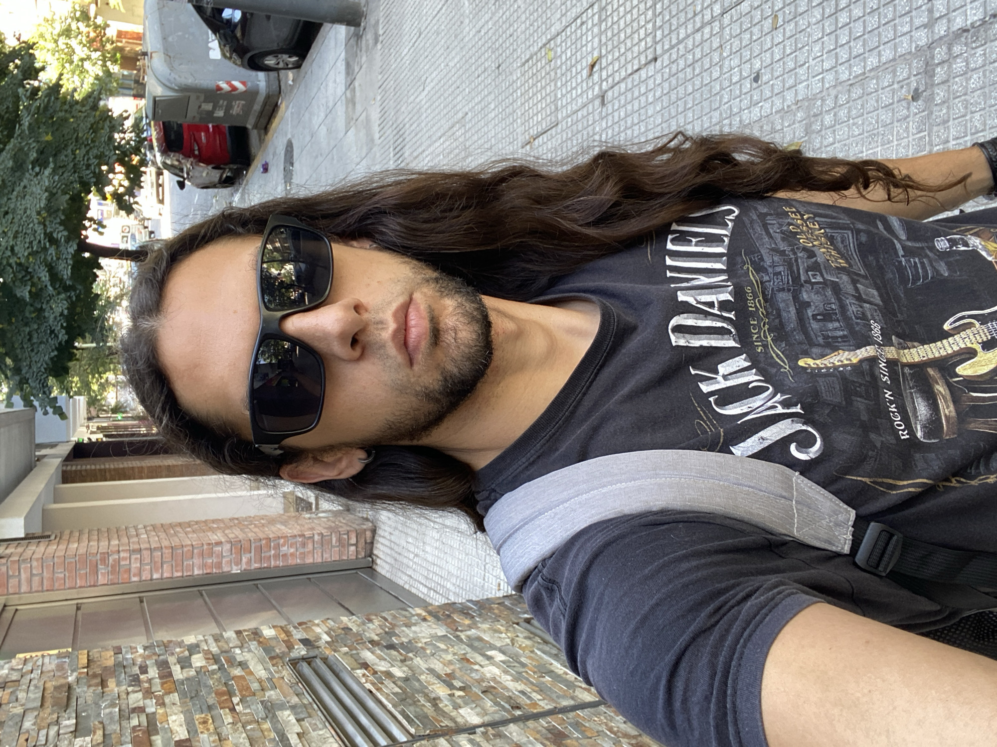 A man is the primary subject of a selfie taken outdoors in what appears to be an urban environment. The man has long, dark brown hair and is wearing sunglasses and a t-shirt with "Jack Daniel's" printed on it. He has a beard and light skin.
The background shows a city street. There are buildings with brick and stone facades, and a paved sidewalk. A tree is visible to the left, and cars are parked along the street. The image appears to be taken in Buenos Aires, Argentina, based on the architectural style and general urban environment.