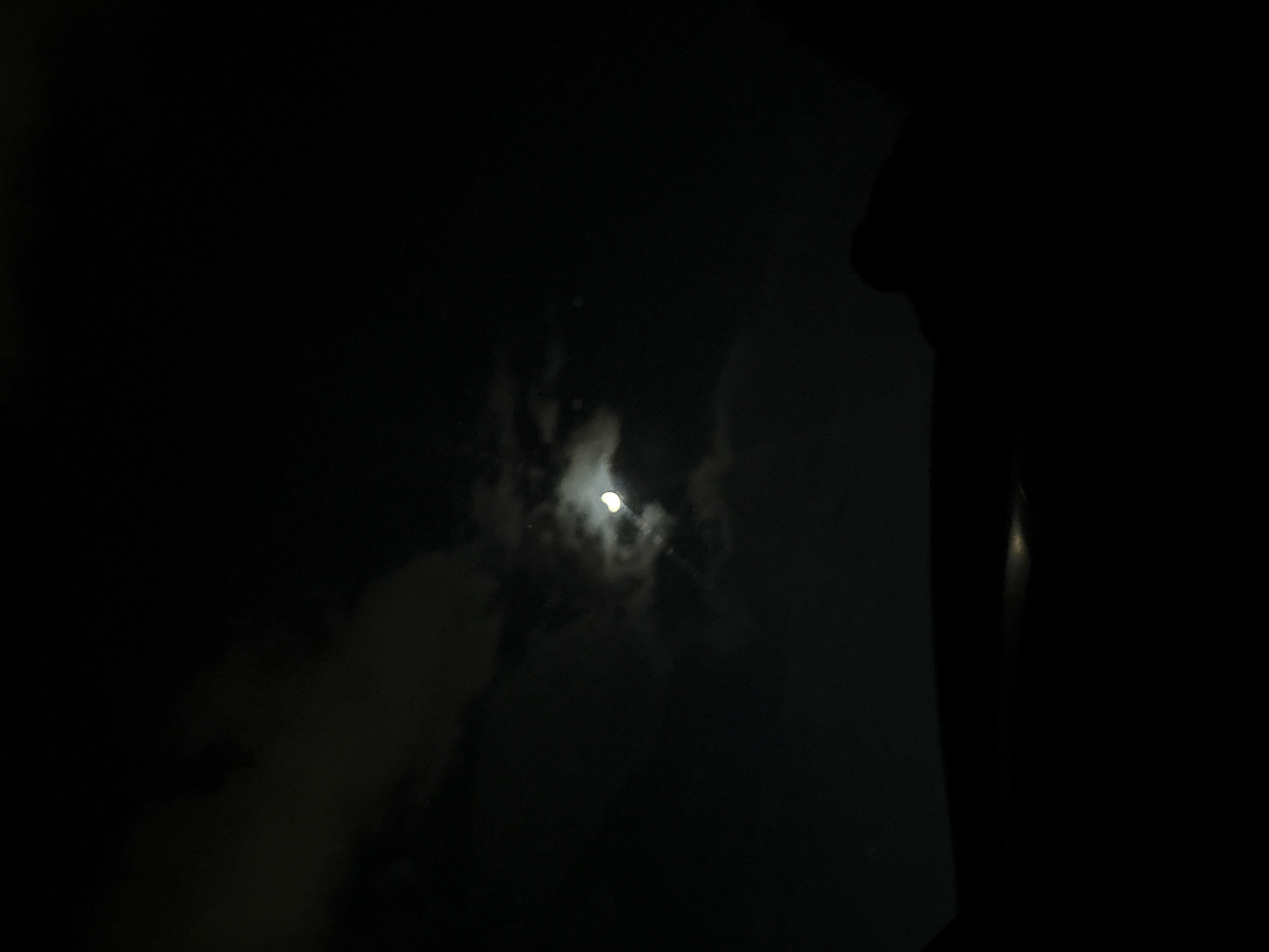 The image depicts a dark night scene. A bright moon is visible, partially obscured by clouds. The surroundings are mostly black, making it difficult to discern details. A dark silhouette is present on the right side of the image. The location in Marulevo, Bulgaria cannot be verified from the image.