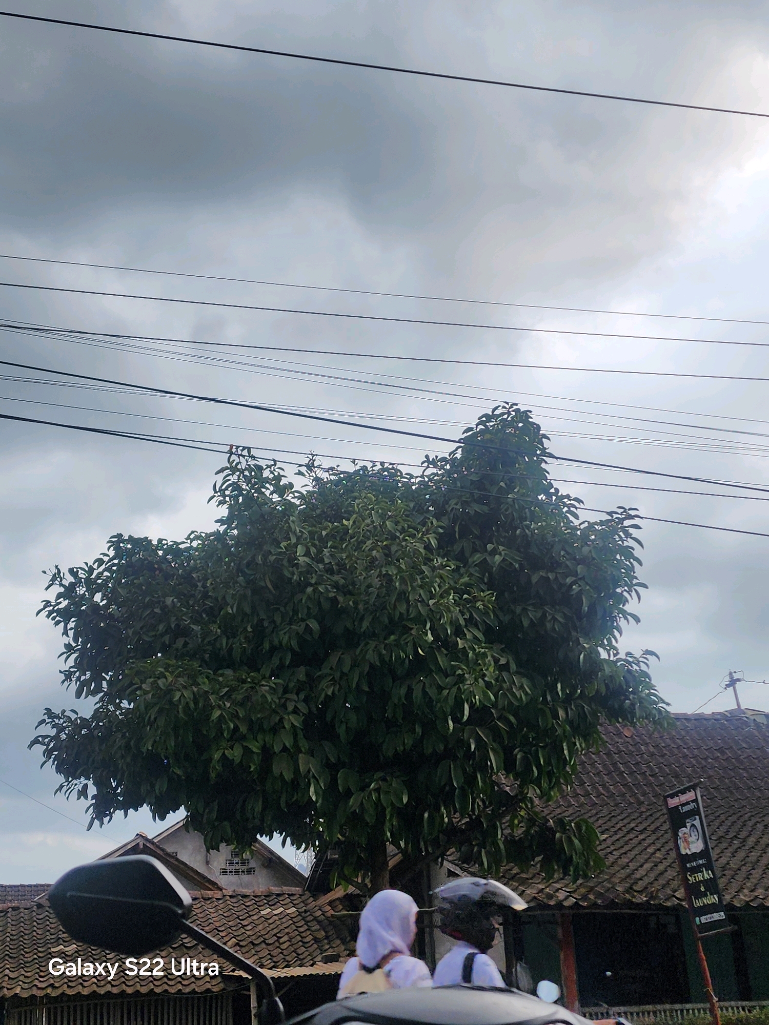 The image shows a street scene in Salaman, Indonesia, under a cloudy sky. In the foreground, a motorcycle side mirror is visible, with the text "Galaxy S22 Ultra" overlaid. Two individuals wearing helmets and headscarves are seated on the motorcycle, facing away from the camera. One person appears to be wearing a white headscarf and white clothing, while the other wears a black helmet and white shirt. Behind them, a large, leafy tree dominates the midground, its branches extending upwards and outwards. Beyond the tree, a tiled roof of a building is visible, and a dark sign with text and images is attached to a pole. Power lines are strung across the sky in the upper portion of the image. The overall impression is of a street on a day with overcast weather, likely in a residential or commercial area of Salaman.