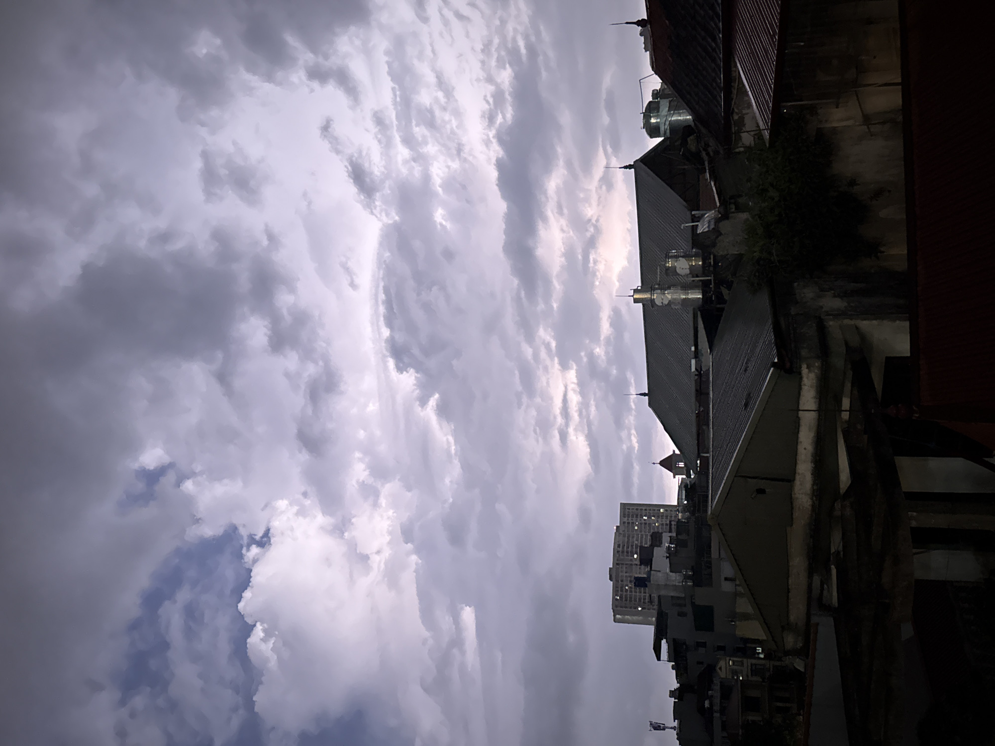 The image shows a view in Hà Nội, Vietnam, captured from an elevated position, possibly a rooftop or high-rise building. The camera is angled upwards, focusing primarily on the sky filled with thick, gray-white clouds that dominate the scene. A small patch of blue sky is visible in the lower-left corner.
Below the clouds, the tops of various buildings are visible, including residential houses with tiled roofs and a taller, modern apartment building in the middle distance. The building exteriors appear aged and weathered, with signs of urban decay. The architecture is a mix of traditional Vietnamese and modern styles.
The lighting suggests it is either late evening or an overcast day. The color palette is muted, with shades of gray, white, and dark tones, contributing to a somber or dramatic mood. There are no people visible in the shot.