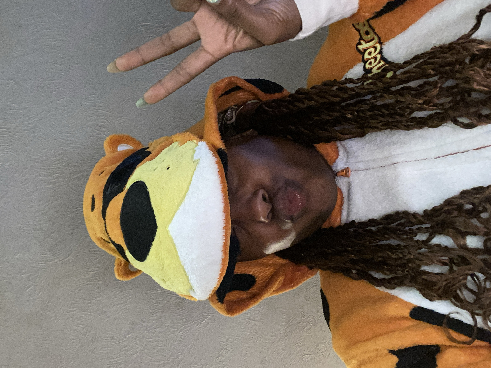 This rotated portrait features an individual wearing an orange, yellow, black, and white kigurumi (animal onesie), specifically themed after Chester Cheetah, the Cheetos mascot. The person has dark skin and long, dark brown braided hair that cascades down their side. Their head is tilted, showing a slightly pouting expression on their face. One hand is raised above their head, forming a peace sign with two fingers; the fingernails are painted in soft, pastel shades of light green and light beige.
The kigurumi's hood features prominent ears and facial elements in orange, yellow, and black. The torso part of the onesie is white, and the brand name "Cheetos" is visibly printed in gold lettering on the front. The background is a plain, light gray textured wall, suggesting an indoor setting. The overall activity is posing for a casual photo, conveying a playful and relaxed mood. While the provided context is London, United Kingdom, there are no specific visual cues within the image to confirm this location.