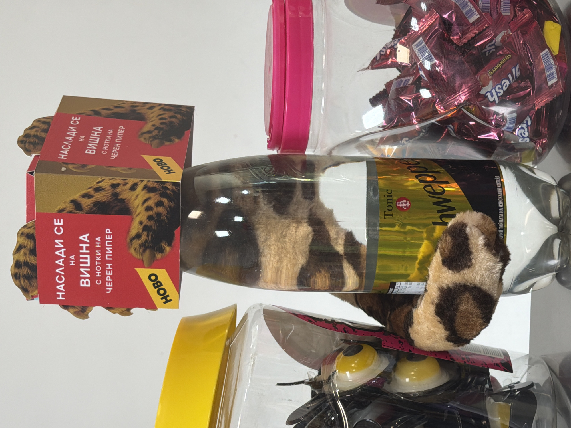 The image shows a close-up view of three plastic containers, one of which is a bottle. The bottle is transparent and contains a fuzzy, leopard-print plush toy arm that appears to be emerging from the top. The bottle has a label with the word "Tonic" and what looks like "Schweppes" written on it. The other two containers are jars, one with a yellow lid and the other with a pink lid. The jar with the yellow lid contains some dark objects with yellow eye-like features. The jar with the pink lid is filled with individually wrapped, red candies. A cardboard box with text in Cyrillic and leopard print is placed on top of the bottle, with parts of the plush toy poking out from behind it. The background is a plain white wall. The scene does not provide any information or visual cues that would suggest a specific location in the city of Dinkata, Bulgaria, or any other specific geographical location.