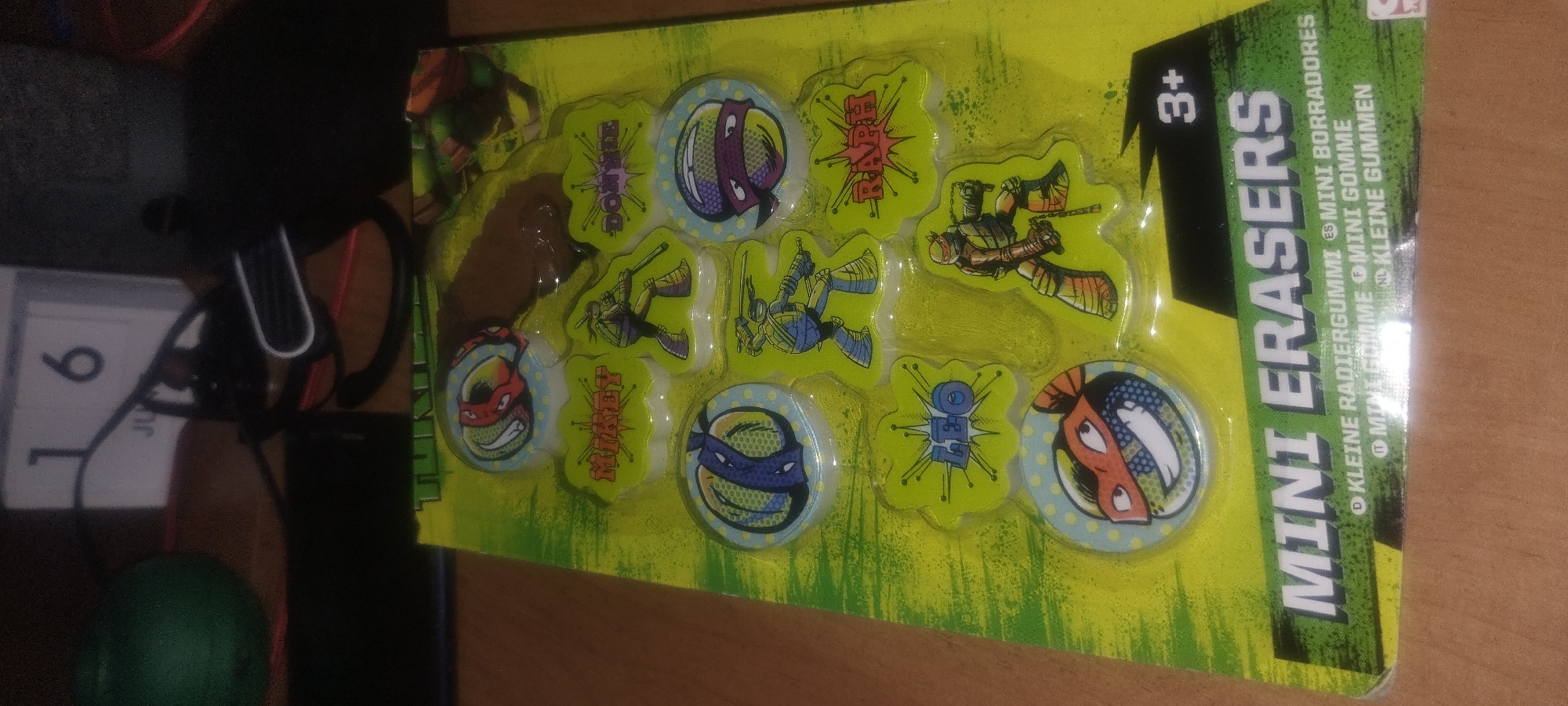 A package of Teenage Mutant Ninja Turtle mini erasers is displayed. The package is primarily green with illustrations of the characters. Each eraser features a character's face or a full-body illustration, along with their name. The erasers are arranged in a grid on the packaging. Some of the names visible are Mikey, Donnie, Leo, and Raph. A "3+" age rating is also present. The packaging appears to be a blister pack, and the erasers are contained within a clear plastic shell. The overall scene is a product shot, likely for retail or promotional purposes.