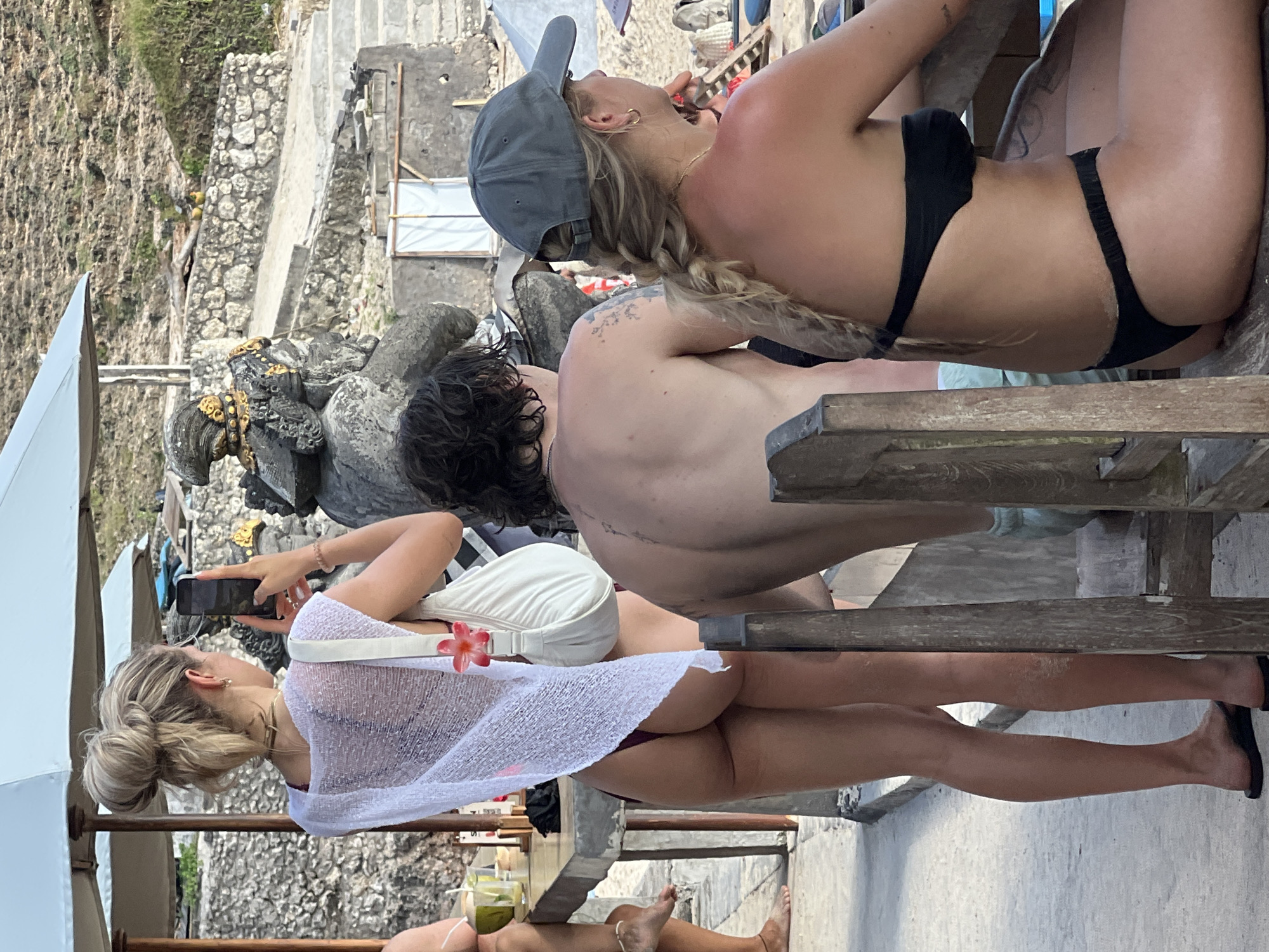 A sunny, outdoor setting in Kuta Selatan, Indonesia shows a group of fair-skinned individuals. At least three of them are seated around a wooden table on a stone patio. One woman is wearing a black bikini and a grey baseball cap with her blonde hair in a braid. A shirtless man with dark hair and a tattoo on his back is seated next to her. Another woman, facing away from the viewer, holds a phone and wears a white cover-up over a bikini, exposing her legs. Stone structures are visible in the background, along with lush foliage and a white sunshade. There is a beverage on the table.