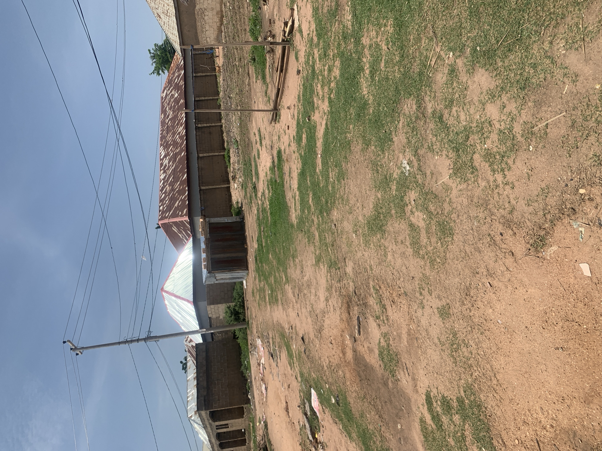 The image shows an outdoor scene in Jauro Yinu, Nigeria, under a clear blue sky. A dirt road with patches of dry grass occupies the foreground. In the middle ground, there are several buildings with corrugated metal roofs, some of which appear rusted. A concrete block wall surrounds some of the properties. A wooden utility pole with electrical wires runs across the upper portion of the image. Some debris, including what looks like discarded paper and plastic, is scattered on the ground. In the background, trees are visible, and there are hints of more structures. The overall impression is one of a rural or semi-urban residential area.