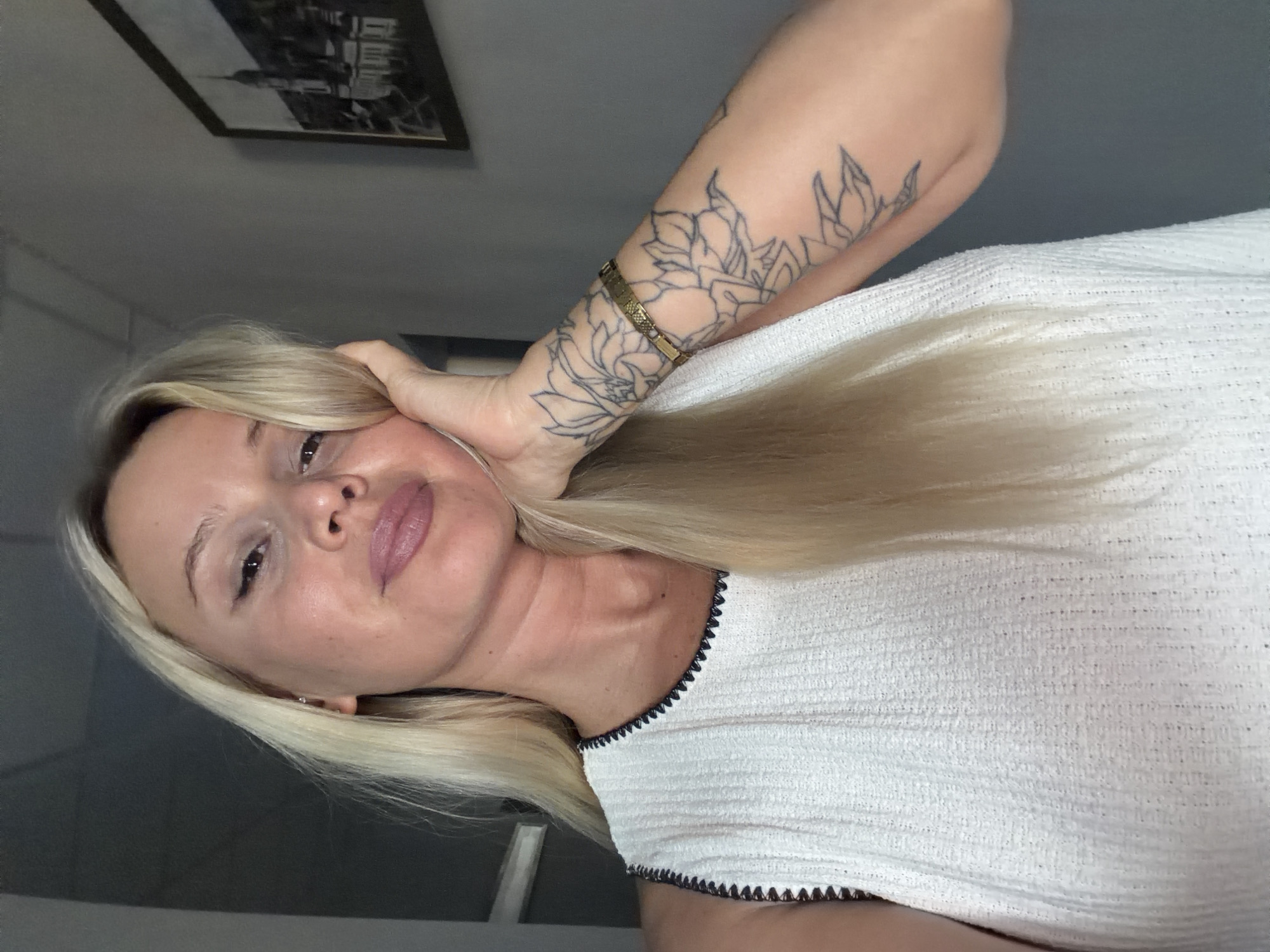 A woman with blonde hair is shown in a close-up, angled shot. She is wearing a white textured top with a dark trim. She has tattoos on her arm, including floral designs. A gold bracelet is visible on her wrist. Her face is turned towards the camera, and her hand is resting on her head. The background appears to be an indoor setting with a framed picture on the wall. The lighting suggests it might be daytime. The woman's expression is neutral. There is no visible text in the image.
