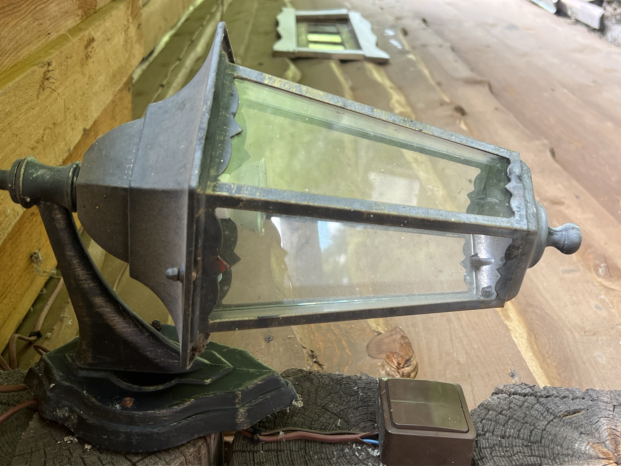 The image captures a close-up of an old, weathered outdoor lantern mounted on a wooden post. The lantern is made of dark metal with glass panels, some of which are dusty. The mounting bracket is ornate and dark, appearing to be attached to a rough wooden surface. Below the lantern, a brown electrical switch with two toggles is visible, connected by wires. The background consists of wooden walls and a yellow ceiling, suggesting an outdoor structure like a porch or eaves. In the far background, a small decorative window with a light-colored frame is visible. The overall impression is one of rustic architecture. The location in Norviliškės, Lithuania, is not discernible from the visual elements present in the image itself.
