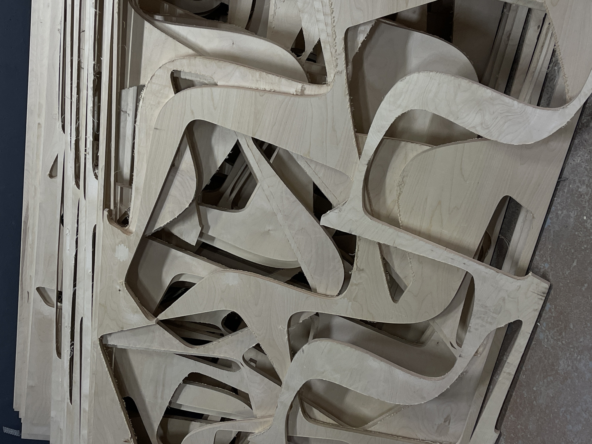 The image depicts a stack of irregularly shaped wooden cutouts, likely from a CNC cutting process, piled on top of each other. The material appears to be plywood, with visible wood grain patterns and rough edges. The cutouts have curved and angular shapes, creating a complex, layered composition. The lighting suggests an indoor environment, possibly a workshop or storage area. The concrete floor is visible in the lower portion of the image. There are no people present in the image. This scene is likely located in Krasnoyarsk, Russia, a city known for its industrial sector and woodworking.