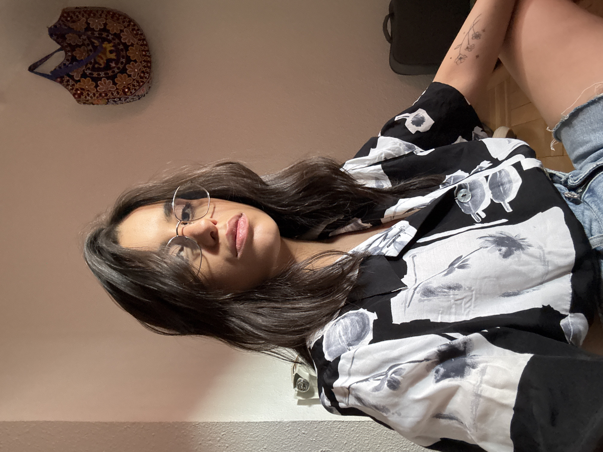A young woman with dark, wavy hair and glasses is seated indoors, looking towards the camera. She is wearing a black and white floral print shirt and denim shorts. A tattoo of a sprig of flowers is visible on her left forearm. Behind her, on the wall, is a colorful, embroidered bag. The room appears to be lit by artificial light, casting shadows. The woman is positioned in the foreground, with the background showing a wall and part of a wooden floor. There is no indication of the location in Las Rozas de Madrid, Spain.
