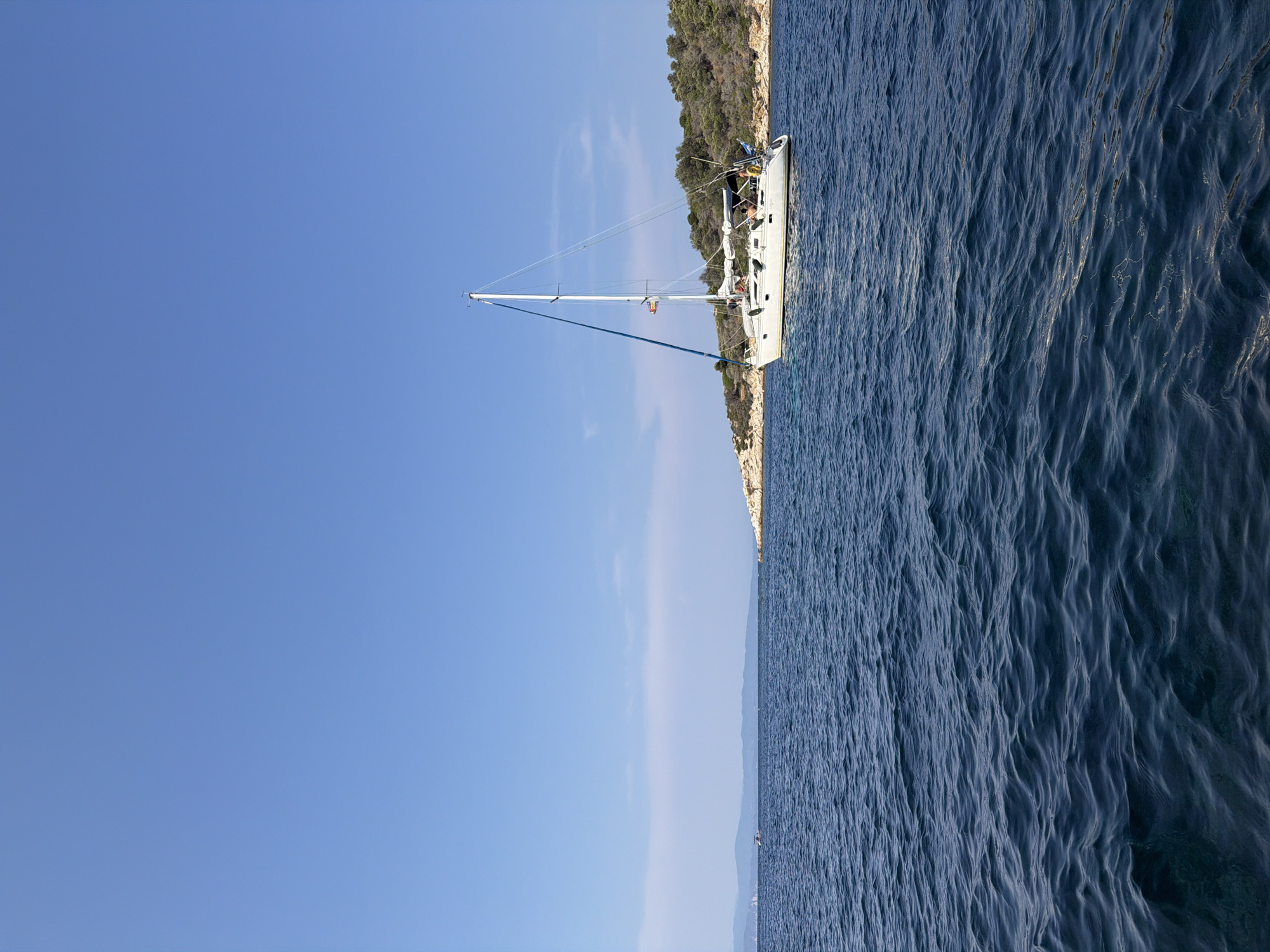 A white sailboat with a blue stripe on its mainsail is moored in the Aegean Sea, off the coast of a rocky, tree-covered island. The boat has several people on board, some appearing to be sitting on the deck. The water is a deep blue, with small waves. The sky is clear and bright blue. In the distance, a hazy coastline is visible. The image was taken from another boat on the water.