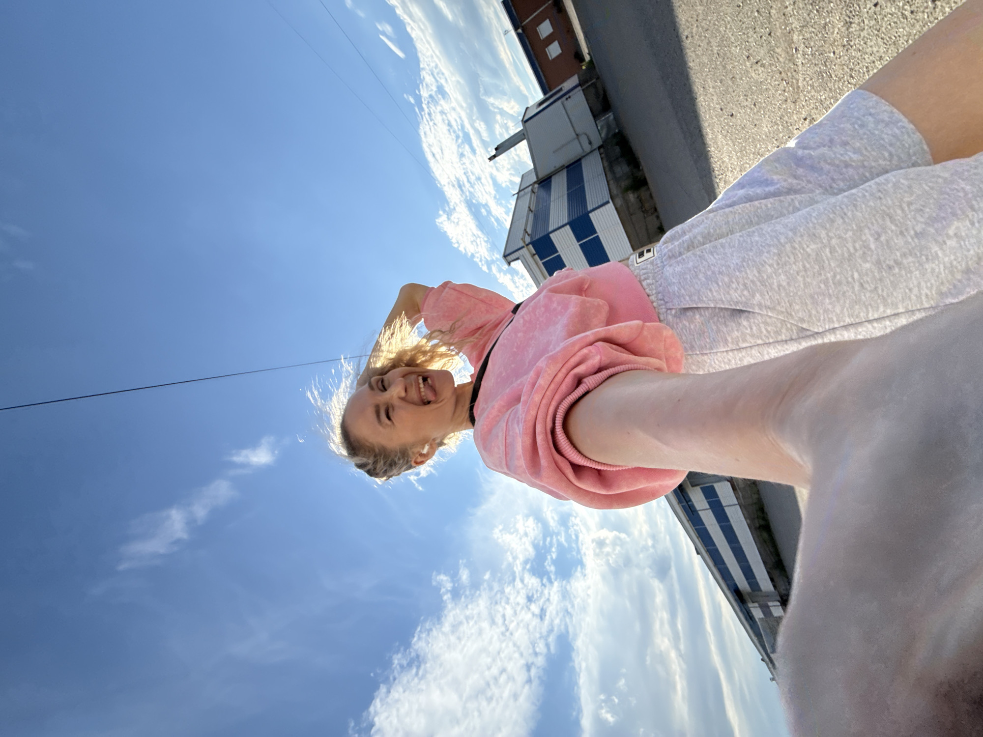 The media file is a low-angle, wide shot of a young woman outdoors, with the sky and a building in the background. The woman is in the foreground, smiling and sticking her tongue out, with her hair falling forward. She is wearing a pink sweatshirt and light gray sweatpants. The scene appears to be a sunny day, as indicated by the bright sky and the clear visibility of the clouds. The setting includes a paved area, possibly a street or sidewalk, and a building with a blue and white striped facade. The building's design suggests an industrial or commercial area. The photo was likely taken in the afternoon, given the quality of the sunlight. The woman's expression conveys playfulness and a sense of joy.