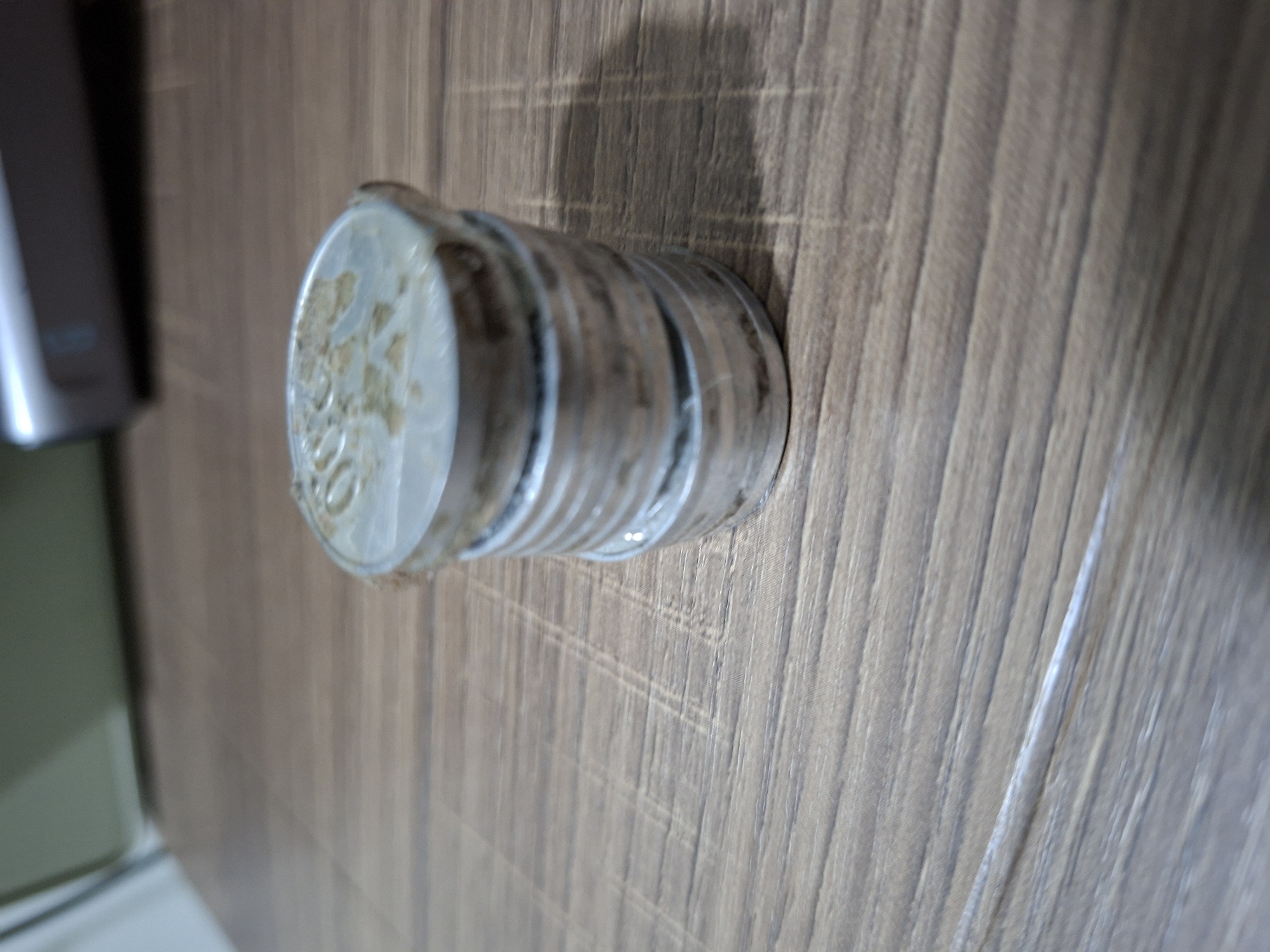 A stack of several metallic coins is placed on a wooden surface. The coins are silver in color and appear to be worn. The wooden surface has a visible grain pattern. In the background, a portion of a silver electronic device is visible. The lighting suggests an indoor setting. The image does not provide any information to determine the location of Samarinda, Indonesia.