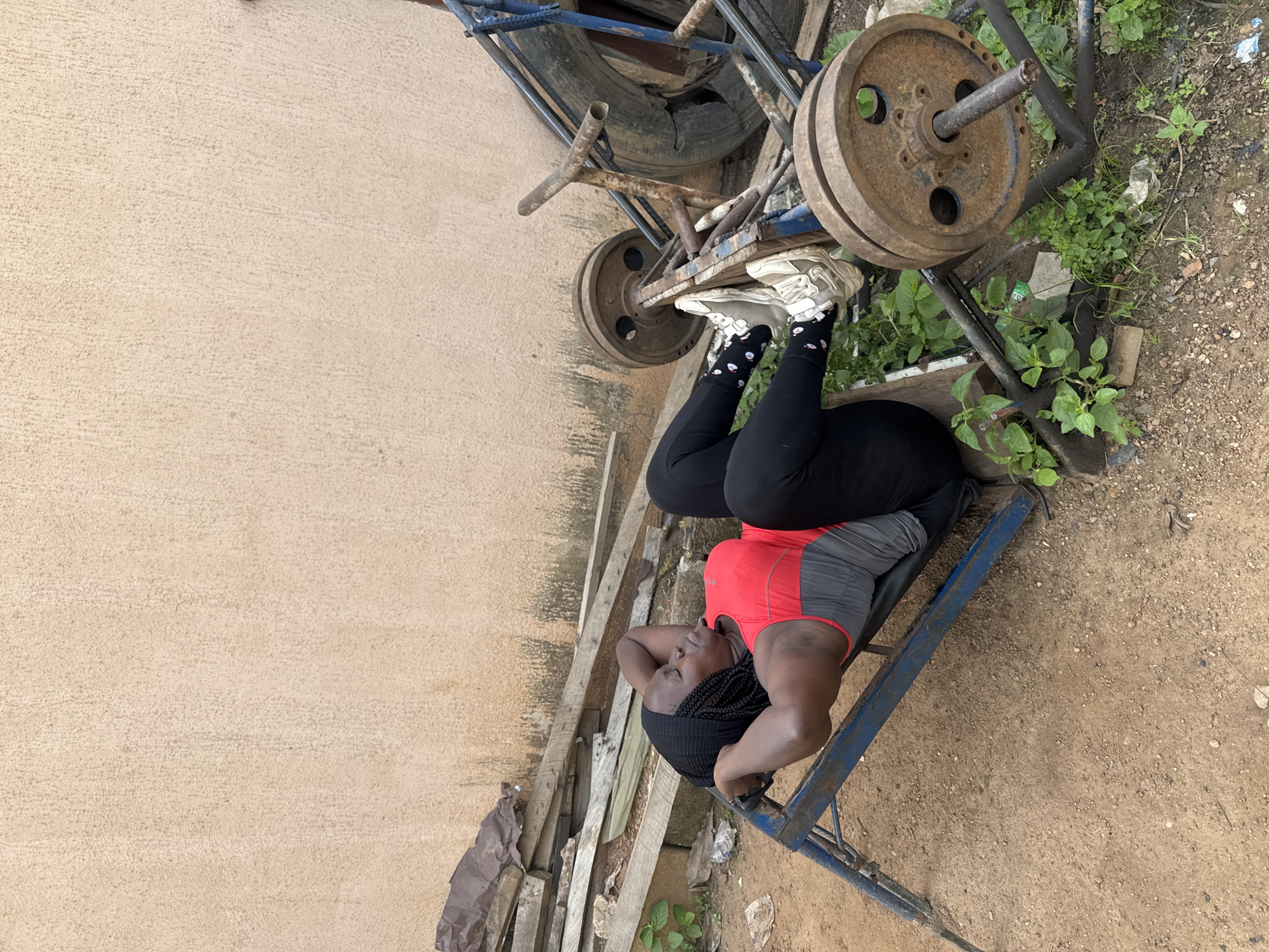 In the city of Akwanga, Nigeria, a Black woman is performing sit-ups on a worn-out blue exercise bench outdoors. She is wearing a black headscarf, a red and grey sleeveless workout top, black leggings, and white sneakers with a black and white patterned sock. Her hands are behind her head. The exercise bench is positioned on a dirt ground next to a beige-colored concrete wall. To the left of the bench, a pile of weathered wooden planks and some debris are visible. To the right of the bench, a rusty weightlifting machine with several heavy circular metal plates and a tire is present. Green weeds and plants are growing around the exercise equipment and on the ground. The scene appears to be an outdoor gym or a makeshift workout area.