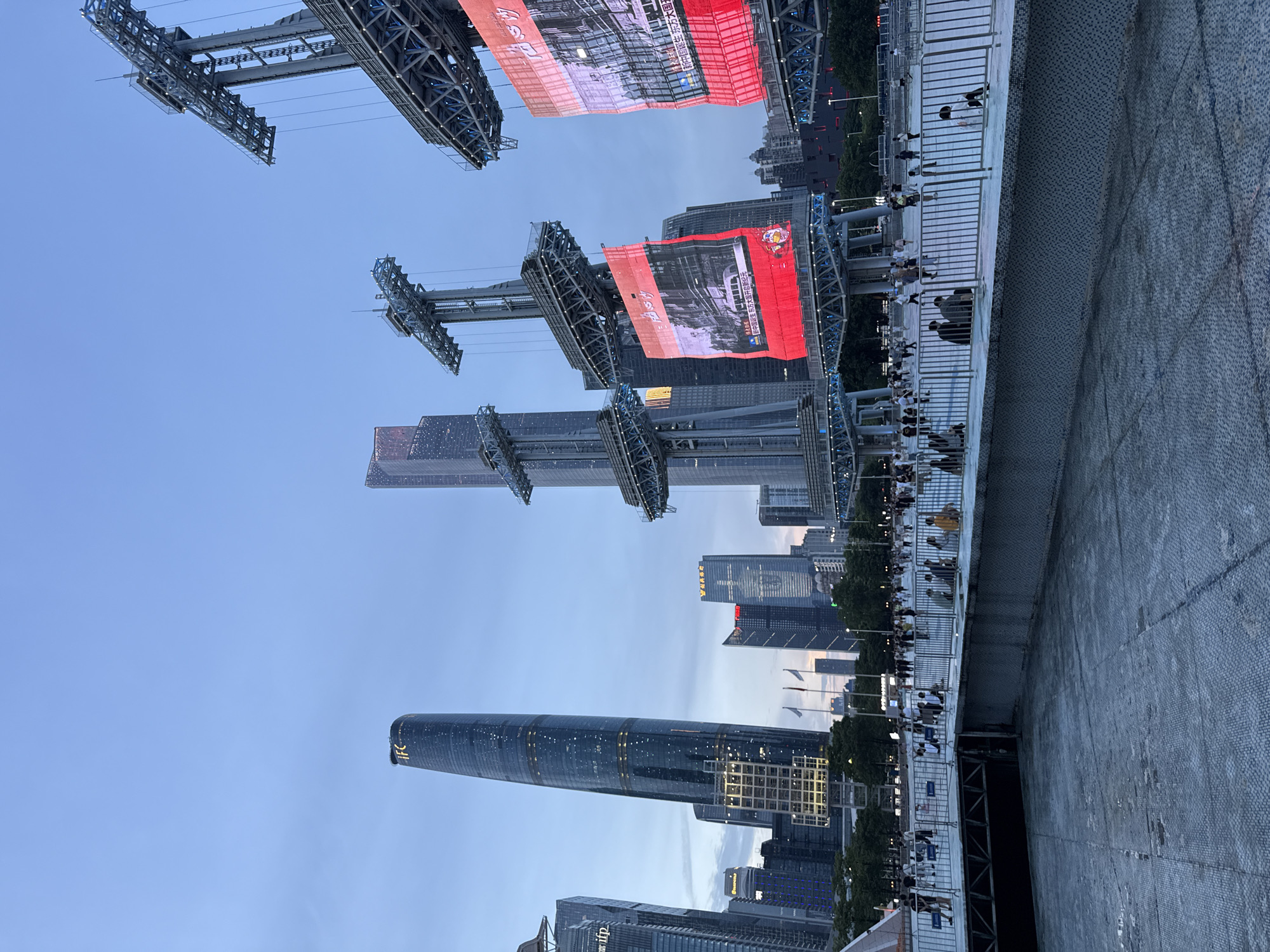 This media file captures an outdoor scene at dusk, likely in a bustling urban area given the presence of skyscrapers. The image is oriented vertically, with the sky at the top and the ground at the bottom. Prominently featured are large, illuminated LED screens suspended from metal structures, displaying advertisements or event information. One screen shows text in Chinese, including "新中国诞生 东方大国开启新纪元" (New China is born, the great Eastern power embarks on a new era) and "新中国诞生 东方大国" (New China is born, the great Eastern power).
Below the screens, a modern cityscape is visible, characterized by tall, glass-clad buildings, including a distinctive cylindrical skyscraper with illuminated floors and the "IFC" logo. A pedestrian walkway with a metal railing runs along the foreground, with numerous people standing and observing. Some are leaning on the railing, while others are further back. The mood appears to be one of anticipation or enjoyment, possibly for an event taking place or a light show. The sky is a soft blue, indicating the transition from day to night, with subtle clouds visible. The overall setting suggests a public gathering space within a major city, likely for entertainment or celebration.