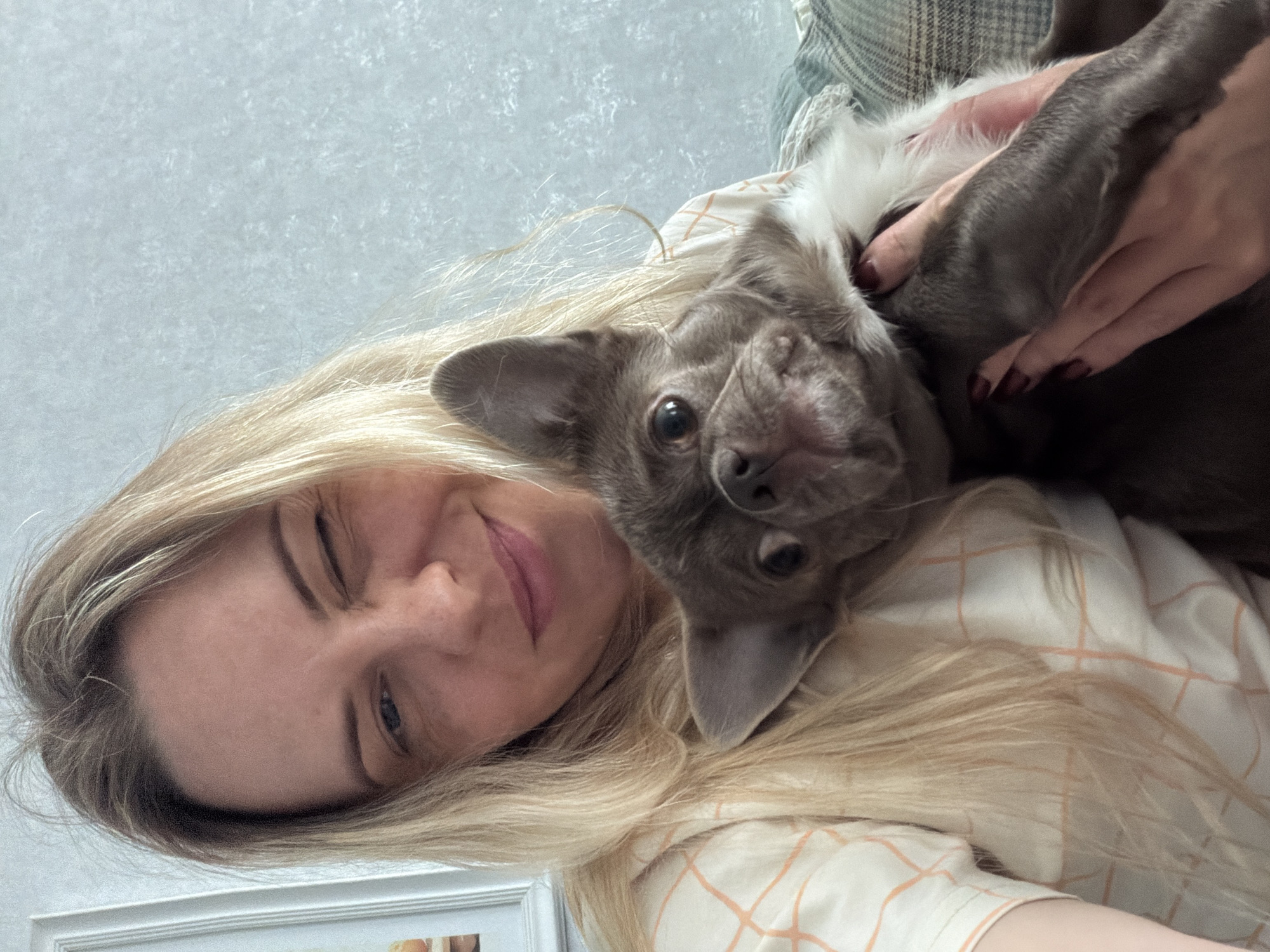 A woman with blonde hair is holding a small, gray and white dog. The woman is winking at the camera. The dog is looking at the camera with its head upside down. The background is a light blue wall. The location is not discernible from the image.
