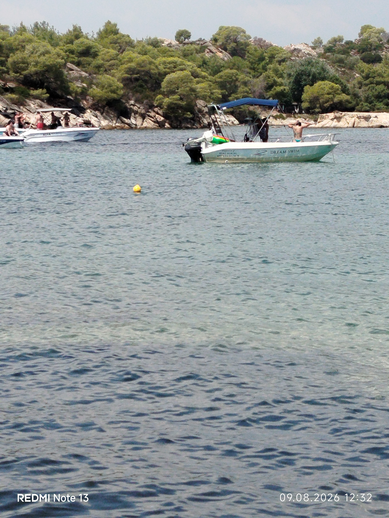 A boat named "DREAM SWIM" is positioned in the water, with people visible on board. A second boat, labeled "ALFA," is to the left, also with individuals on it. The boats are in the sea, with a rocky, tree-covered coastline in the background. The location is identified as Sithonia Municipal Unit, Greece. The sky is overcast, and the water is rippled. A yellow buoy is floating in the water. The timestamp "09.08.2026 12:32" and "REDMI Note 13" are visible at the bottom of the image.