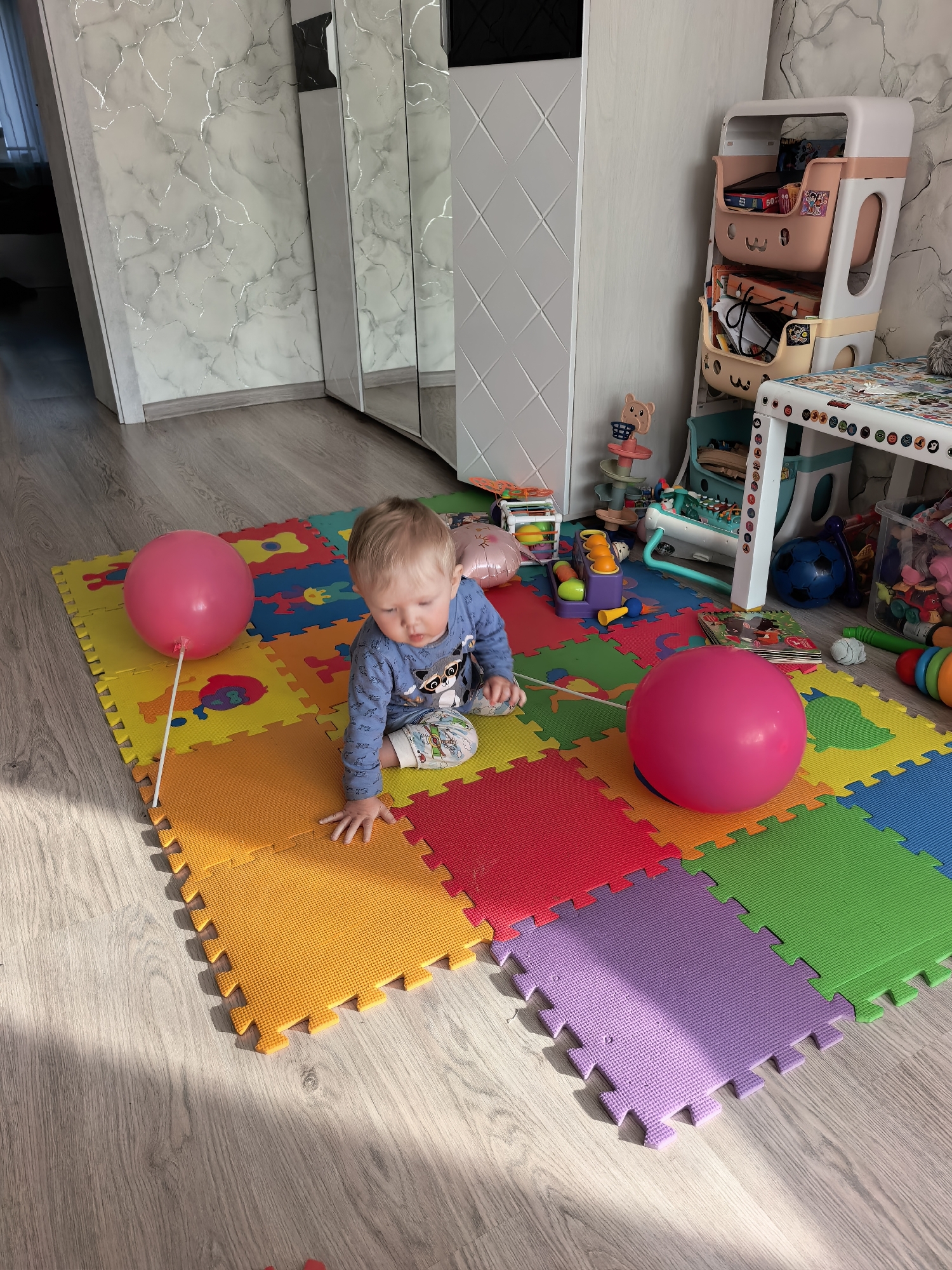 This media file captures an indoor scene featuring a baby in a home located in Sterlitamak, Russia.
**Subjects:** The primary subject is a young baby, appearing to be a toddler, with light blonde hair, dressed in a blue long-sleeved top featuring a cartoon raccoon, and light-colored pants.
**Objects:** The baby is positioned on a vibrant, multi-colored foam play mat composed of interlocking square tiles in shades of red, orange, yellow, green, blue, and purple. Two pink balloons are visible: one large, spherical balloon resting on the mat, and a smaller, oval-shaped foil balloon partially visible behind the baby. The room is filled with various toys, including a clear plastic storage bin, a white play table with colorful designs on its surface, a tiered toy storage unit with pink and beige bins, a blue soccer ball, a colorful toy ramp with balls, and other plastic play items. The flooring is light wood-patterned laminate. A wall features light grey wallpaper with distinct silver, marble-like patterns, and a large mirrored wardrobe or cabinet reflects parts of the room.
**Activity or event taking place:** The baby is in a crawling or kneeling position on the play mat, looking down, seemingly focused on an object just out of frame or the mat itself. The overall impression is one of a child at play in a designated area.
**Notable details:** Natural sunlight streams into the room from an unseen window, casting bright highlights and distinct shadows on the floor and play mat, indicating it is daytime, possibly morning or late afternoon. The environment is clearly set up for a young child, emphasizing play and safety. The baby's expression is one of calm focus.
**Visible text:** The number "60Z" is faintly visible on one of the pink toy storage bins. The white play table's edge has small, circular, colorful decorative stickers or elements that may contain text or numbers, but they are not clearly legible.
