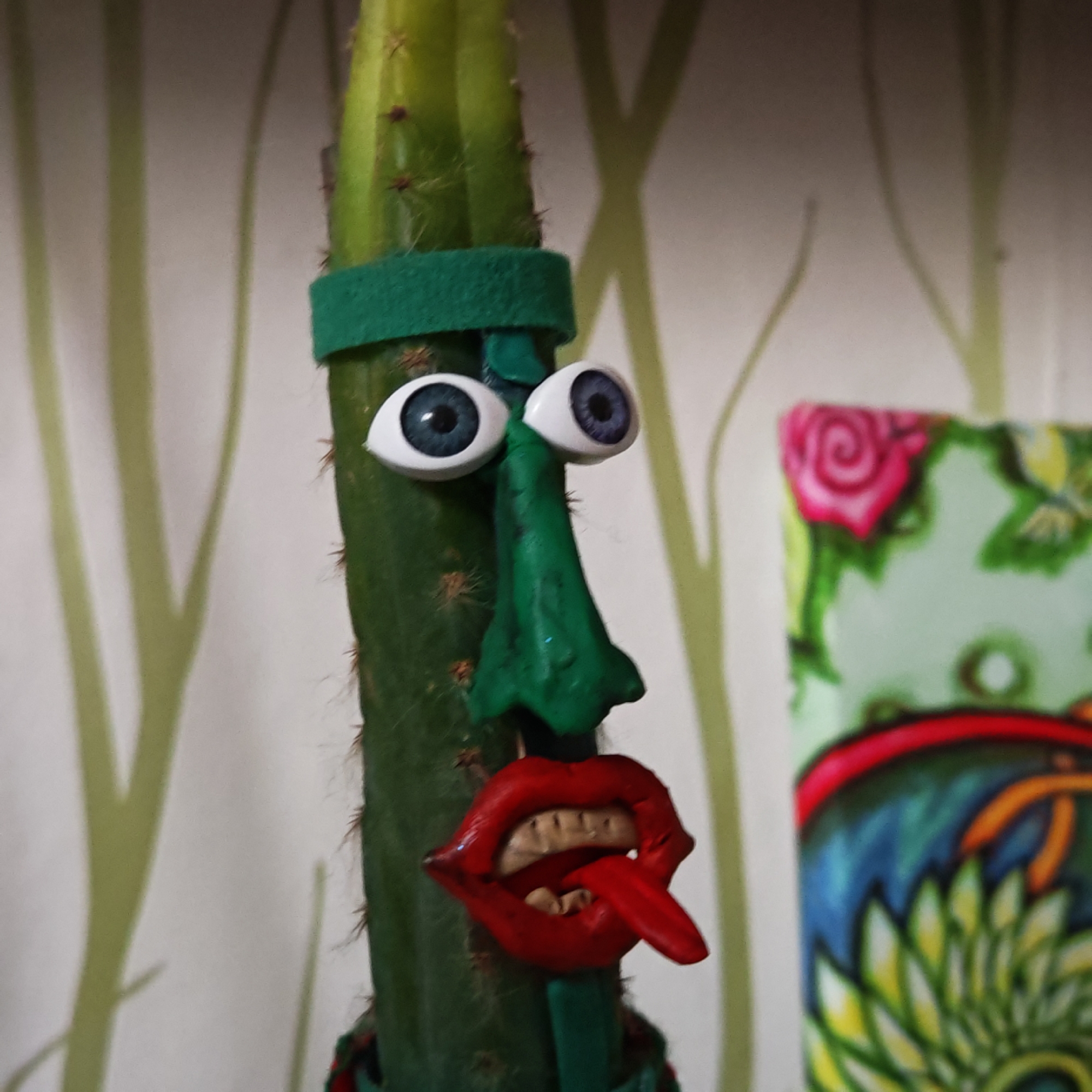 A close-up shot shows a cactus decorated with a whimsical face. The cactus itself is tall and green with small spines. It's adorned with large, cartoonish googly eyes, a long, green, clay-like nose, and a pair of bright red, exaggerated lips with teeth and a protruding tongue. A green felt headband encircles the top portion of the cactus, and another green felt ring is visible near the base. The backdrop is a light-colored wall with a stylized tree branch pattern. To the right, slightly out of focus, is a decorative object featuring bright floral and abstract designs. The image was taken in Novo-Nikol'skoe, Russia.
