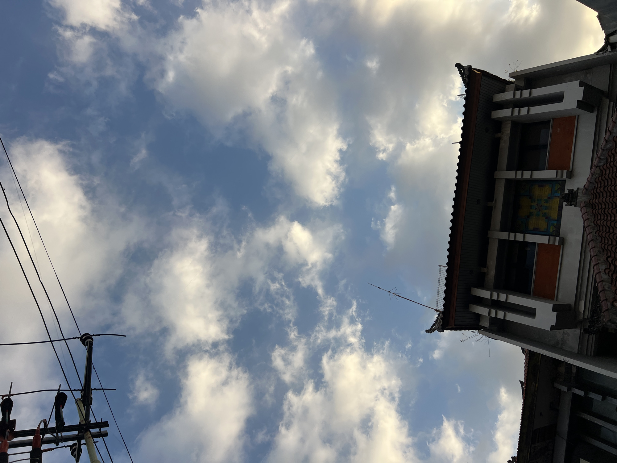 In Kuta, Indonesia, an outdoor shot shows a close-up, low-angle view of the sky, a utility pole and wires, and the upper portion of a building. The sky is a clear blue with scattered white clouds. The utility pole is black with several wires extending from it, some of which are taut and straight, while others are slack. The building has a traditional tiled roof and an ornately decorated facade. The scene is illuminated by natural light, suggesting it was taken during the day.