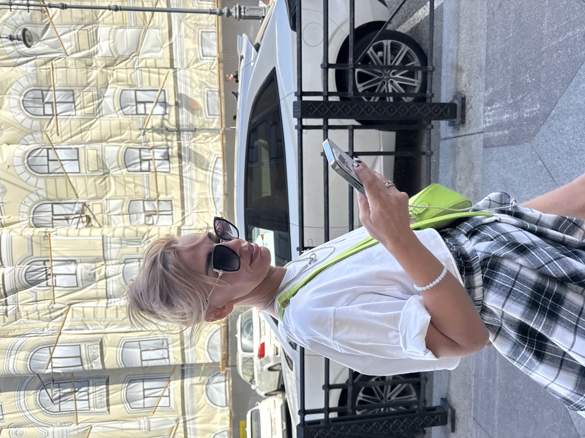 In Saint Petersburg, Russia, a blonde woman with her hair in a bun, wearing a white t-shirt, a plaid skirt, and sunglasses, walks while looking at her phone. She has a green purse slung over her shoulder and wears jewelry on her neck and wrist. The background features a building covered in a protective tarp, with scaffolding around it, and several cars parked on the street, including a white SUV.
