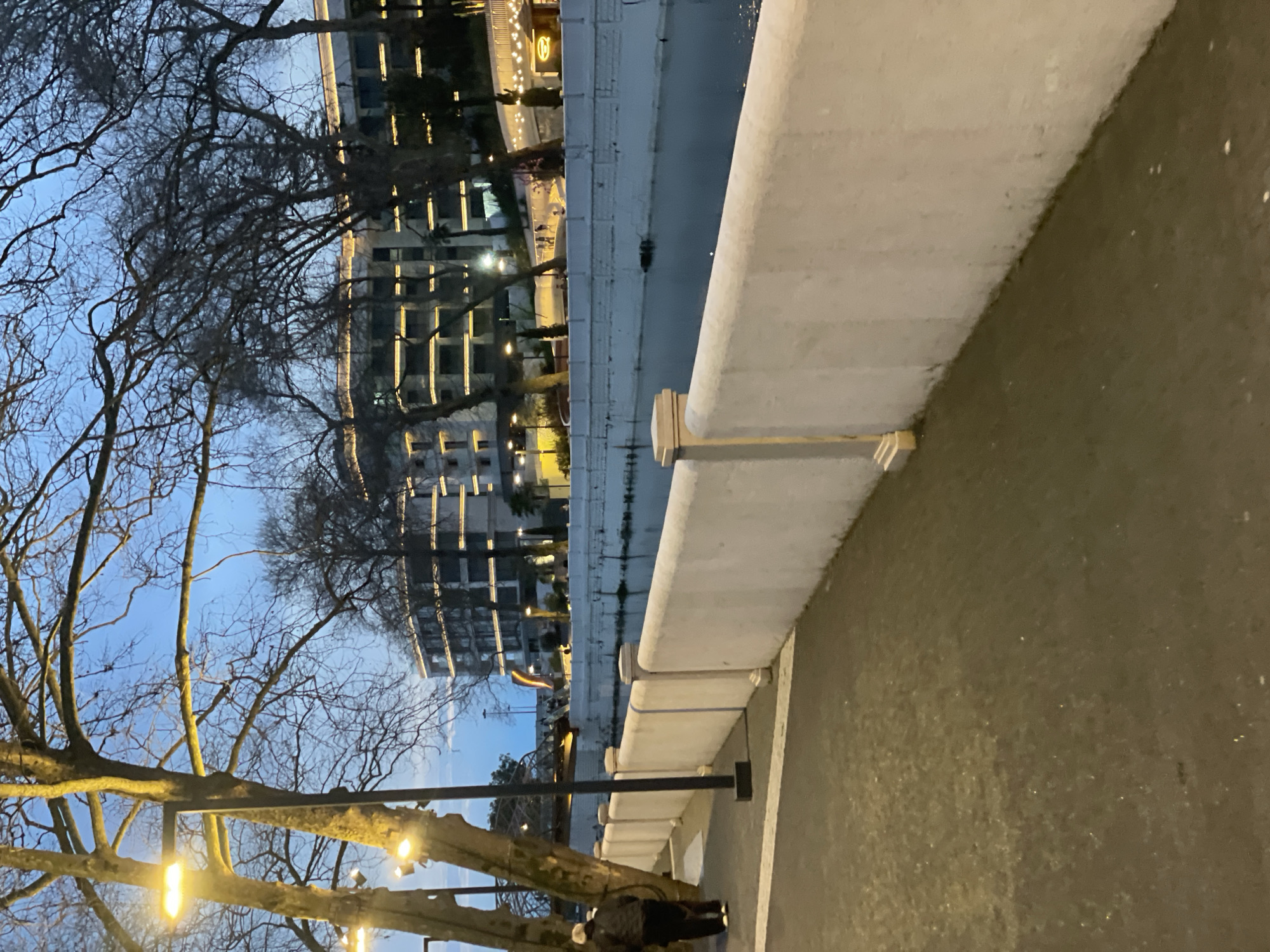 The image presents a view from a pedestrian area alongside a river in what appears to be an urban setting. Buildings line the opposite bank of the river, displaying illuminated windows that suggest either residential or commercial occupancy. The architecture seems modern with multiple stories. A concrete retaining wall or barrier stands between the pedestrian walkway and the river. A bare tree with lights attached to its branches is visible in the foreground, suggesting a decorative or festive lighting scheme. The sky suggests dusk or early evening. A person is standing by the bare tree. Based on the architecture and geography, this appears to be in Sochi, Russia.