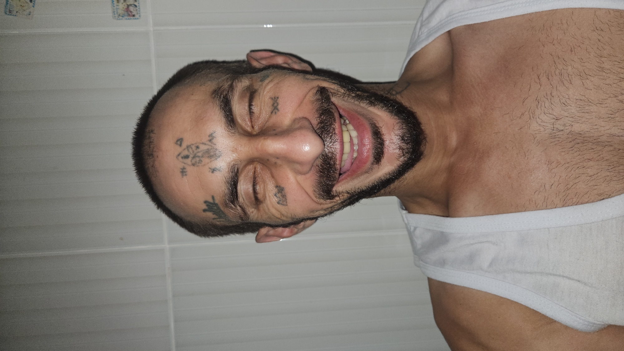 The image displays a close-up portrait of an adult male, captured indoors in Cam Đức, Vietnam. The man is depicted smiling broadly with his eyes closed, conveying a sense of happiness or amusement. He has a dark, full beard and mustache, along with very short, almost shaved hair on his head.
His face is notable for several distinct tattoos. Centered on his forehead is a stylized design, possibly a fish or an animal, with a small symbol resembling the number "4" positioned above and to its right. A prominent "XX" mark is tattooed on his right cheekbone, while a more abstract, darker symbol, potentially a leaf or flame, is visible on his left cheekbone. Another small, dark mark appears under his left eye.
He is wearing a plain white tank top, part of which is visible over his chest and shoulder. The background consists of a white tiled wall featuring horizontal grout lines. Faint, possibly decorative, old stickers or small pictures can be seen on the upper part of the wall. The lighting appears even and artificial. The entire image is oriented sideways, rotated 90 degrees counter-clockwise.