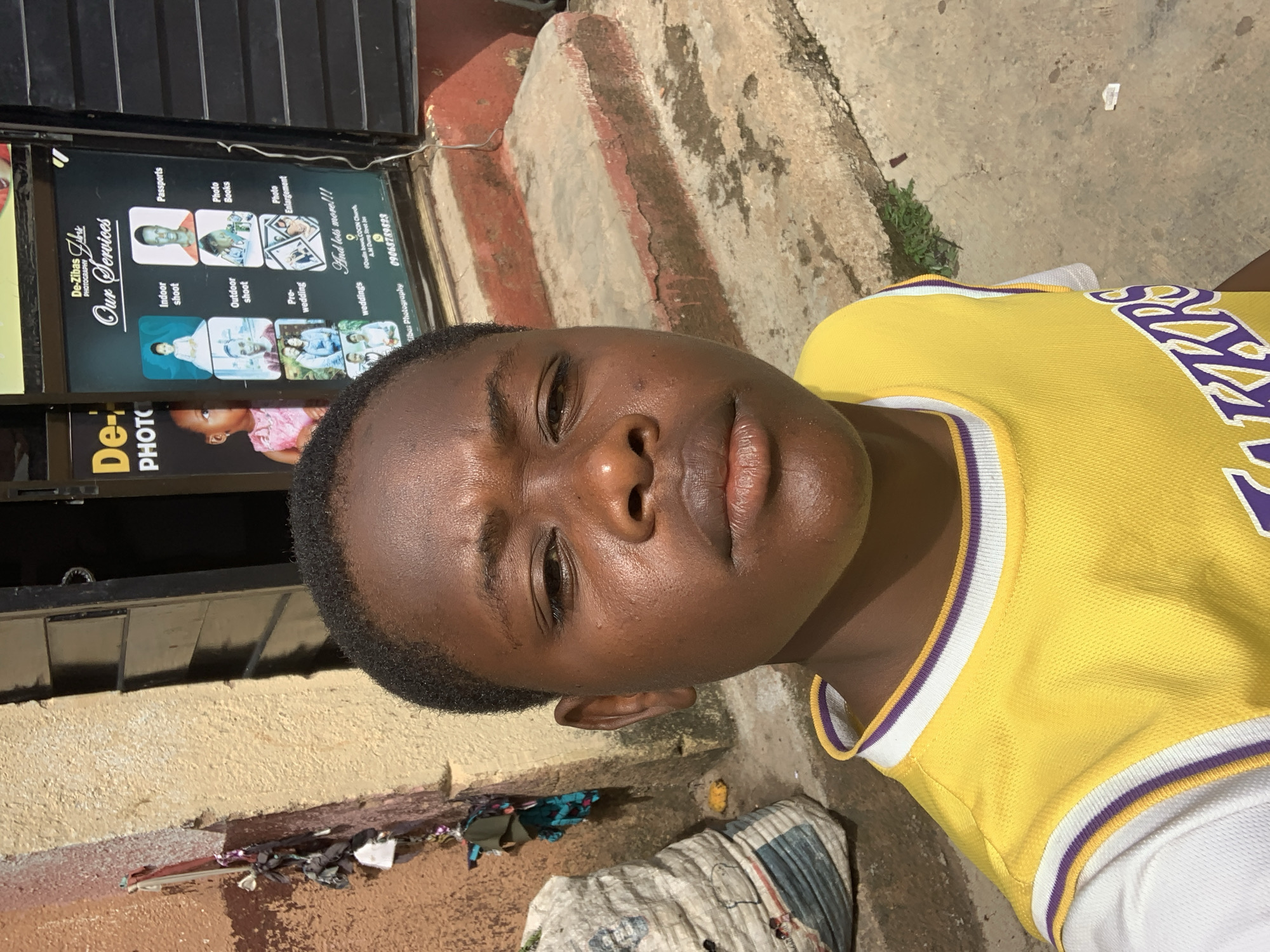 In Jos, Nigeria, a young boy with dark skin and short black hair takes a selfie in front of a photography studio. He is wearing a yellow Lakers basketball jersey over a white t-shirt. Behind him, a sign for "De-Zibas Photography" lists various services including "Indoor shoot," "Outdoor shoot," "Pre-wedding," "Weddings," "Passports," "Photo Books," and "Photo Enlargement." The studio appears to be a small shop with a dark door and a concrete floor. To the left of the studio, a large, white, woven bag is leaning against a textured, light-brown wall adorned with hanging clothes and some miscellaneous items. The ground is made of concrete, and there are red painted steps leading up to the studio entrance. The scene is bathed in natural daylight, casting shadows and highlighting the textures of the environment.