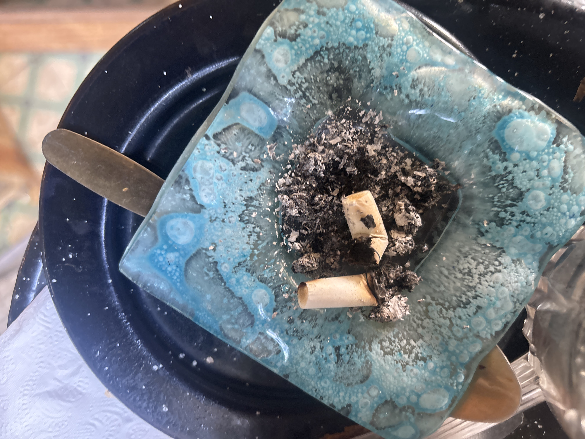 An overhead shot captures a square, glass ashtray filled with ashes and two cigarette butts. The ashtray is made of light blue or teal glass with a distinctive, bubbly, textured surface, particularly along its edges and bottom, which gives it a somewhat organic or water-like appearance. A pile of dark grey and white ash occupies the center of the ashtray, where two light-colored cigarette butts, one of which appears broken, are resting.
The ashtray sits on a dark, possibly black or very dark blue, metallic plate or shallow bowl. This plate has a slightly textured or speckled surface, with small white dots scattered across it. A slender, golden-bronze colored metallic object, resembling a stir stick or a narrow spoon, is partially visible, tucked between the ashtray and the dark plate.
In the bottom left corner, a crumpled white paper towel or napkin is partially visible beneath the dark plate. The background is largely out of focus, but hints of light-colored, patterned floor tiles can be seen in the upper left, suggesting an indoor or covered outdoor setting. The scene does not show any people or active events, but the contents of the ashtray indicate recent smoking. There is no visible text. The location is Buenos Aires, Argentina, though no specific visual cues confirm this.