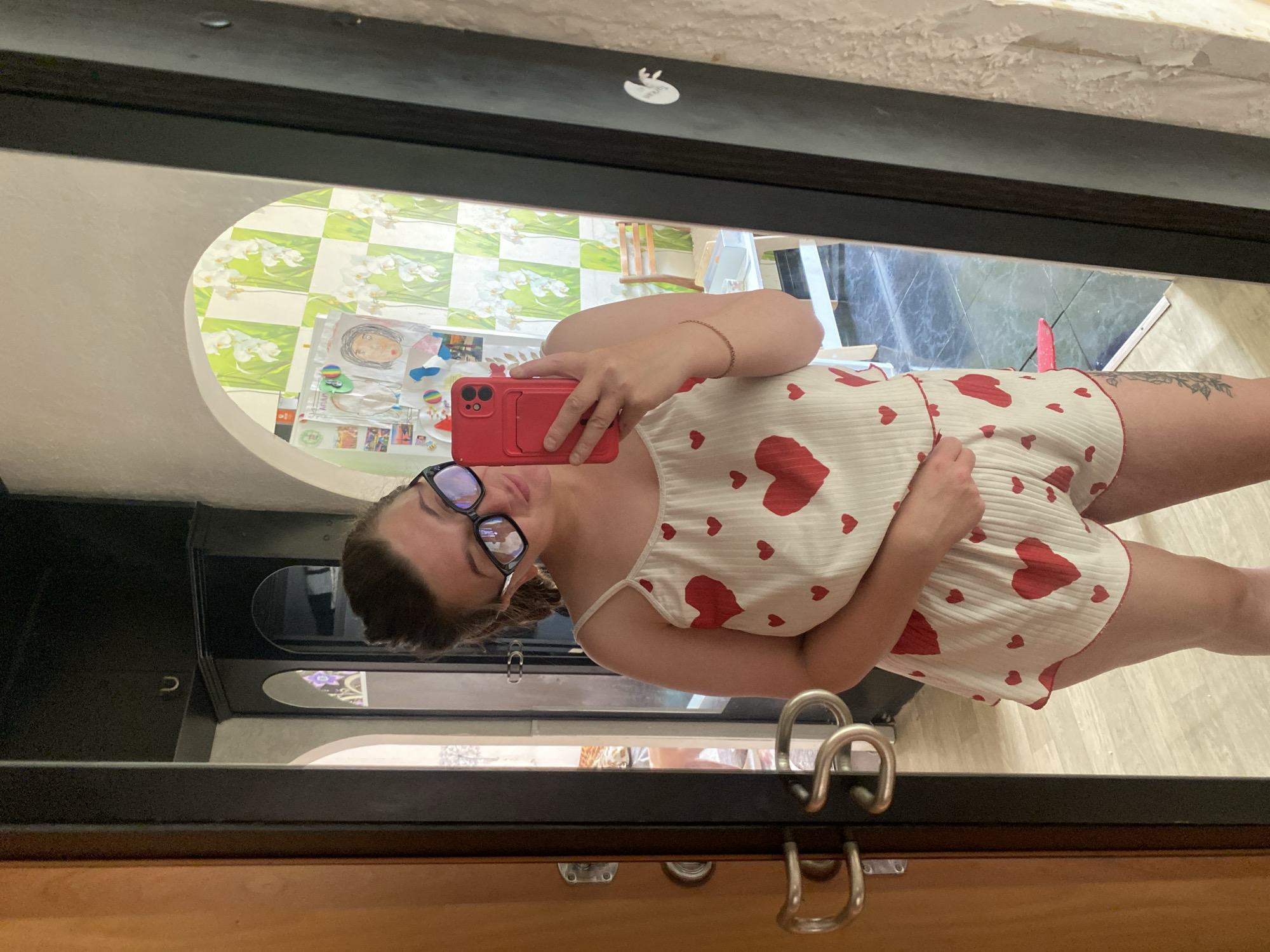 A young woman with dark hair pulled up in a bun, wearing large-rimmed glasses, takes a selfie in a mirror. She is dressed in a white pajama set with a pattern of red hearts, consisting of a camisole and shorts. She holds a red smartphone in her right hand. The background reflected in the mirror shows a wall with a green and white checkered wallpaper adorned with white flowers, and several drawings pinned to it. To the left, a wooden door with metal handles is visible, and beyond it, another room with a bed can be seen. The floor is tiled with a marble pattern. The image was taken indoors, likely in a residential setting, potentially in Chaikovsky, Russia, given the location context provided.