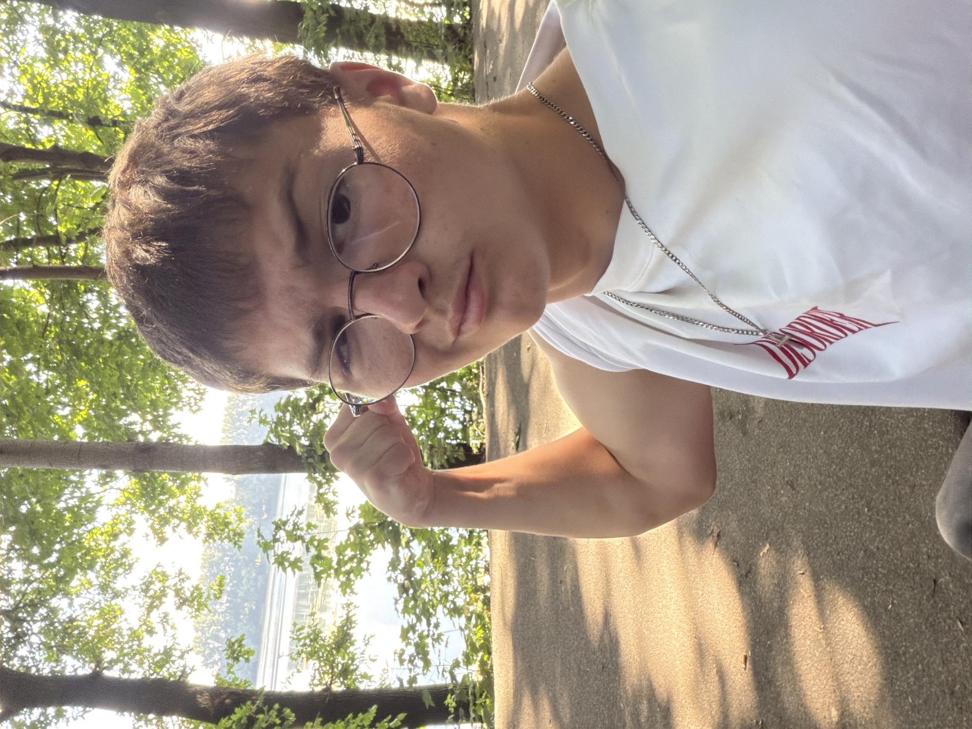 A young person with short brown hair and round glasses is posing for a selfie outdoors, with a body of water and trees in the background. They are wearing a white t-shirt with a red design and a silver chain necklace. The person is flexing their bicep. The scene appears to be a park or recreational area, potentially near the Lippe river in Dorsten, Germany, given the presence of water and trees, and the lighting suggests it is late afternoon or early morning.