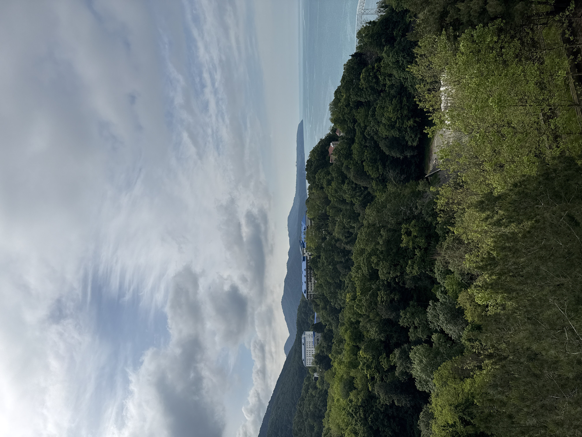 The image depicts a scenic overlook of the Black Sea coastline in Nebug, Russia. In the foreground, a dense, verdant forest covers rolling hills. The forest transitions to a scattering of buildings with blue roofs, appearing to be resort accommodations, nestled amidst the trees on the hillsides. In the mid-ground, the calm, turquoise waters of the Black Sea stretch out towards the horizon, meeting a sky filled with scattered clouds. A white bridge or pier structure is partially visible on the right side of the image, extending over the water. The overall scene conveys a sense of natural beauty and peacefulness, with human structures integrated into the landscape.