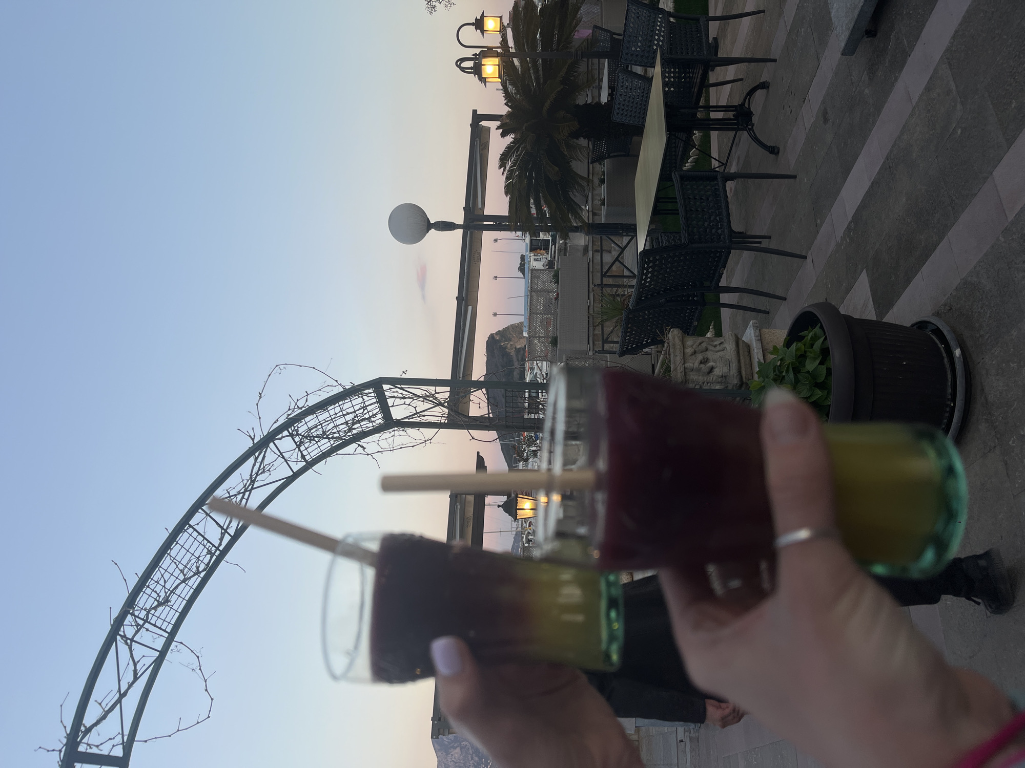 A low-angle shot depicts two cocktails held in hands against the backdrop of an outdoor seating area in Budva, Montenegro, likely near the seaside promenade.
The cocktails are in clear glasses with visible layers of different colors; the top portion appears to be a dark purplish-red, while the bottom is a greenish-yellow. Each has a wooden stick or straw extending from the top. The hands holding the drinks appear fair-skinned, with a silver ring visible on one of the fingers.
In the background, there’s an outdoor restaurant or bar area with black metal chairs and tables. A palm tree and decorative lampposts are visible, suggesting a seaside location. The sky above the scene transitions from a light blue to a pale orange hue, indicating either sunrise or sunset.
An arched trellis structure is seen silhouetted on the left, adding depth to the scene. In the far background, there is a glimpse of the Montenegrin coastline, possibly the Adriatic Sea. The flooring is paved with large gray tiles, suggesting a public area or terrace. A potted plant is present on the right side of the image.